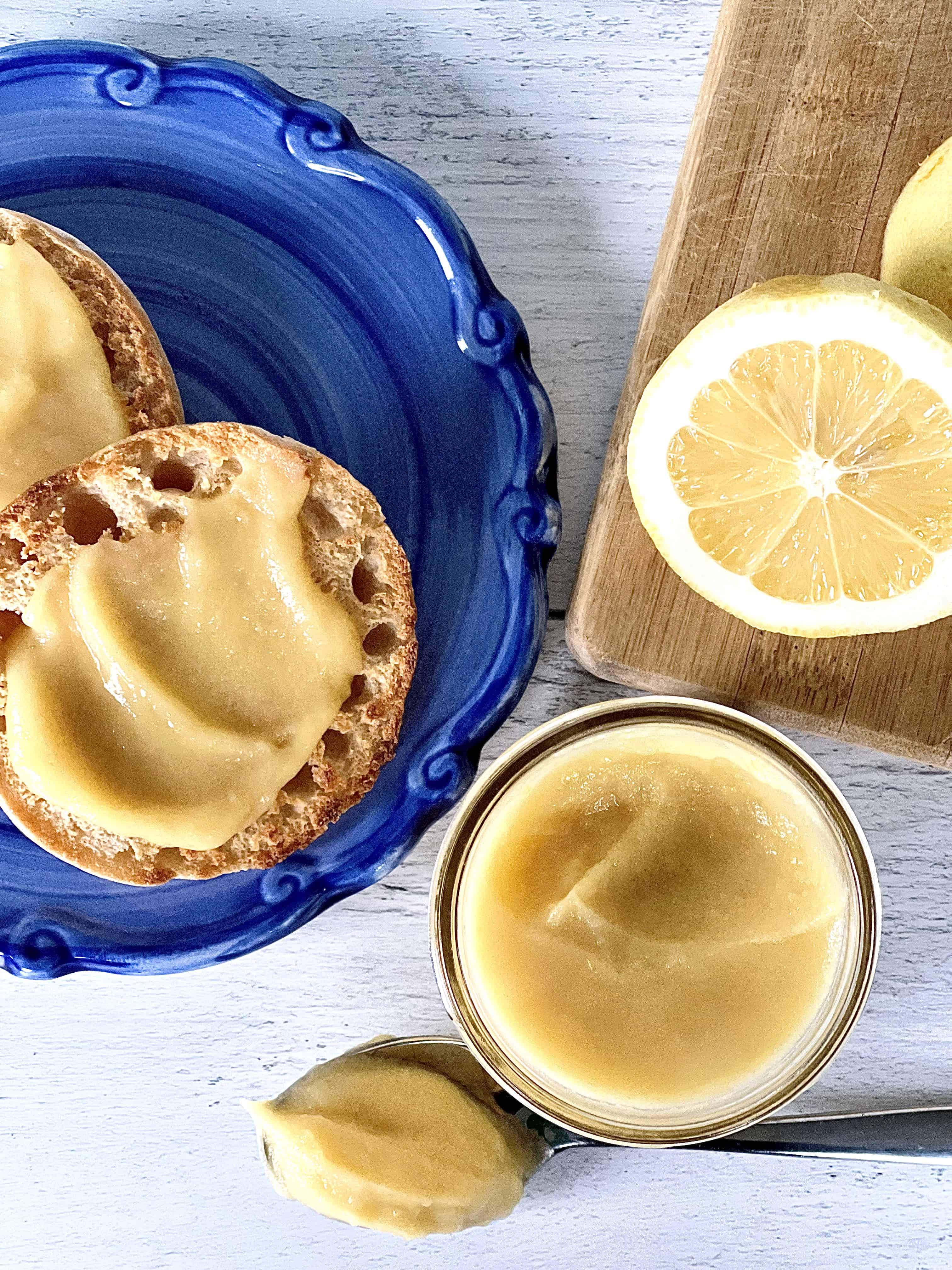 honey lemon curd in a mason jar, on a spoon and on two English muffin halves on a blue swirl plate next to a wooden cutting board with two lemon halves on it