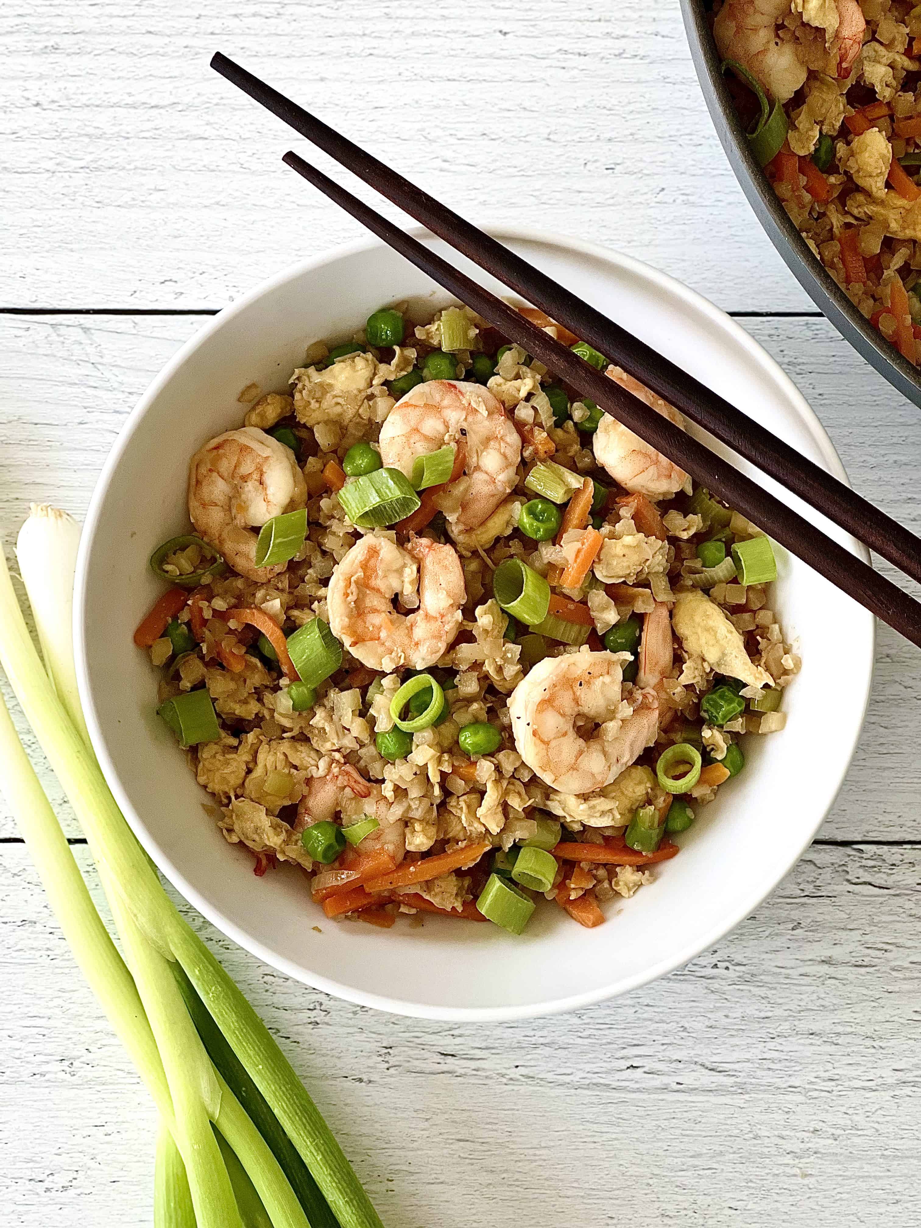 healthy shrimp and rice in a white bowl with chopsticks resting on the rim, next to 3 scallions on a white wooden table