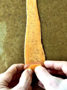 2 hands holding a cocktail sausage as they wrap it up in a strip of sweet potato on a brown cutting board