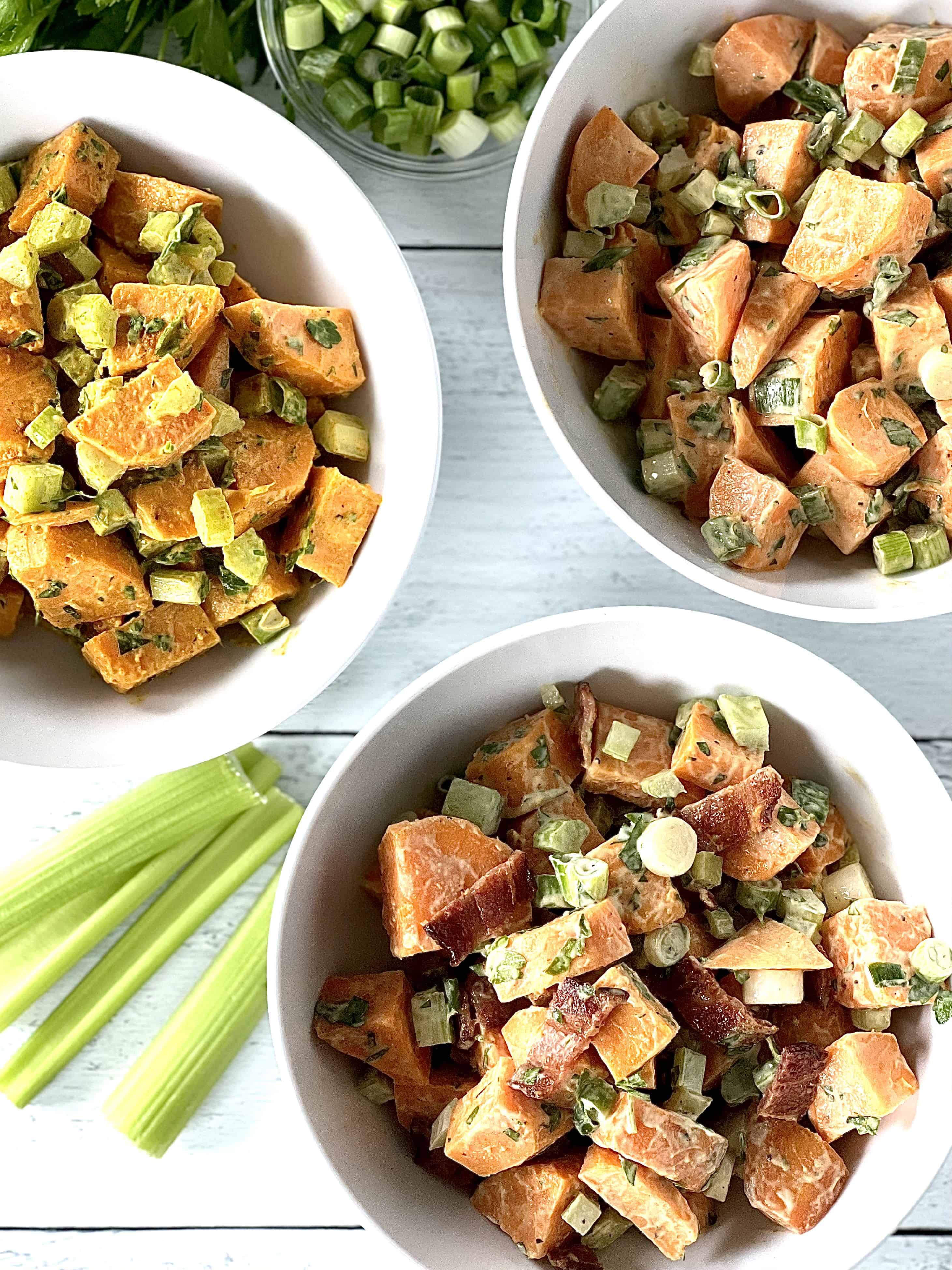 healthy sweet potato salad in 3 white bowls on a white wooden table with celery stalks and a bowl of sliced scallions