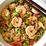 Whole30 Shrimp stir fry in a white bowl with chopsticks resting on the rim, next to 3 scallions on a white wooden table