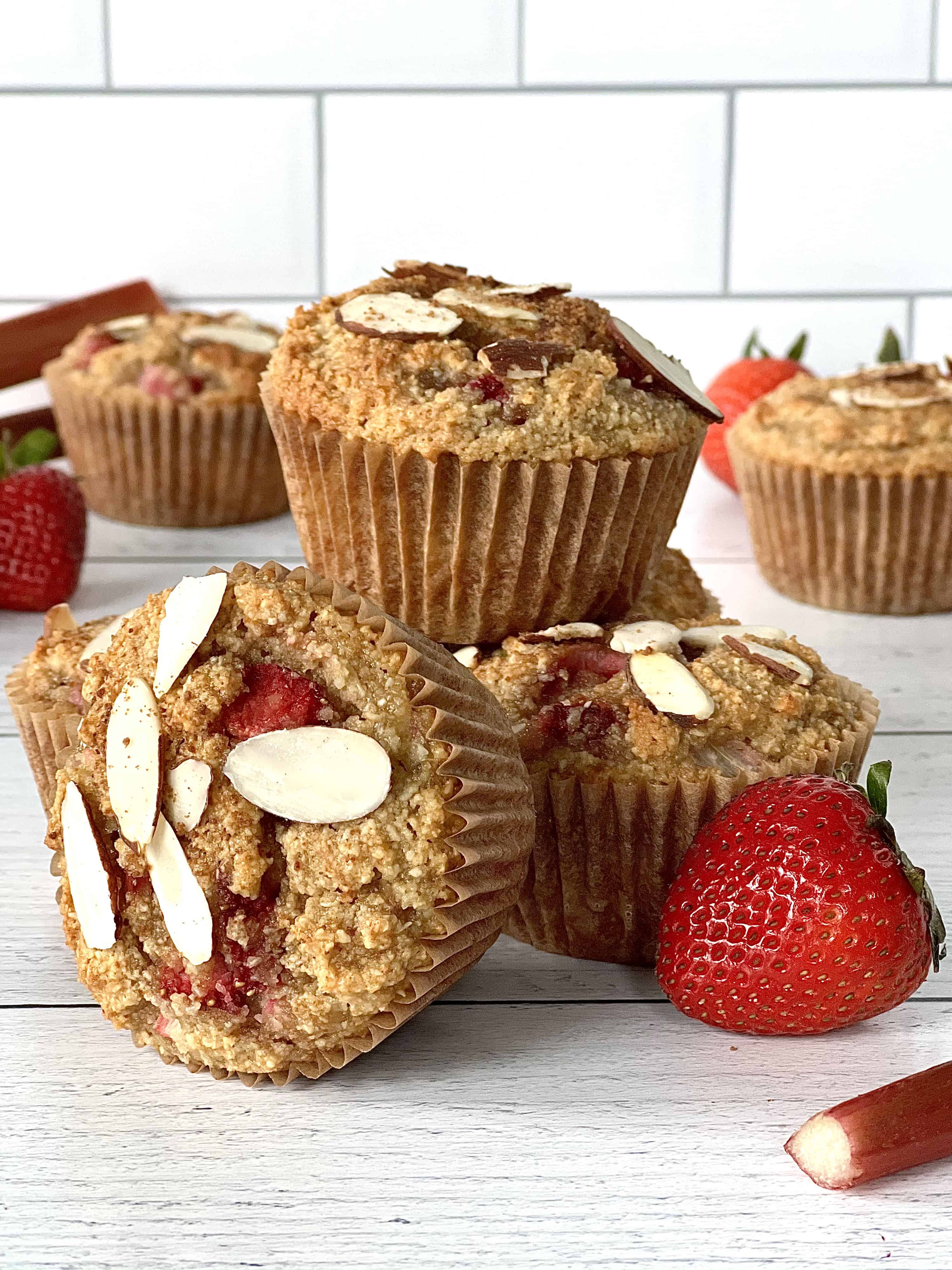 healthy muffins with rhubarb and strawberries in them, topped with sliced almonds, on a white wooden table with rhubarb stalks and strawberries