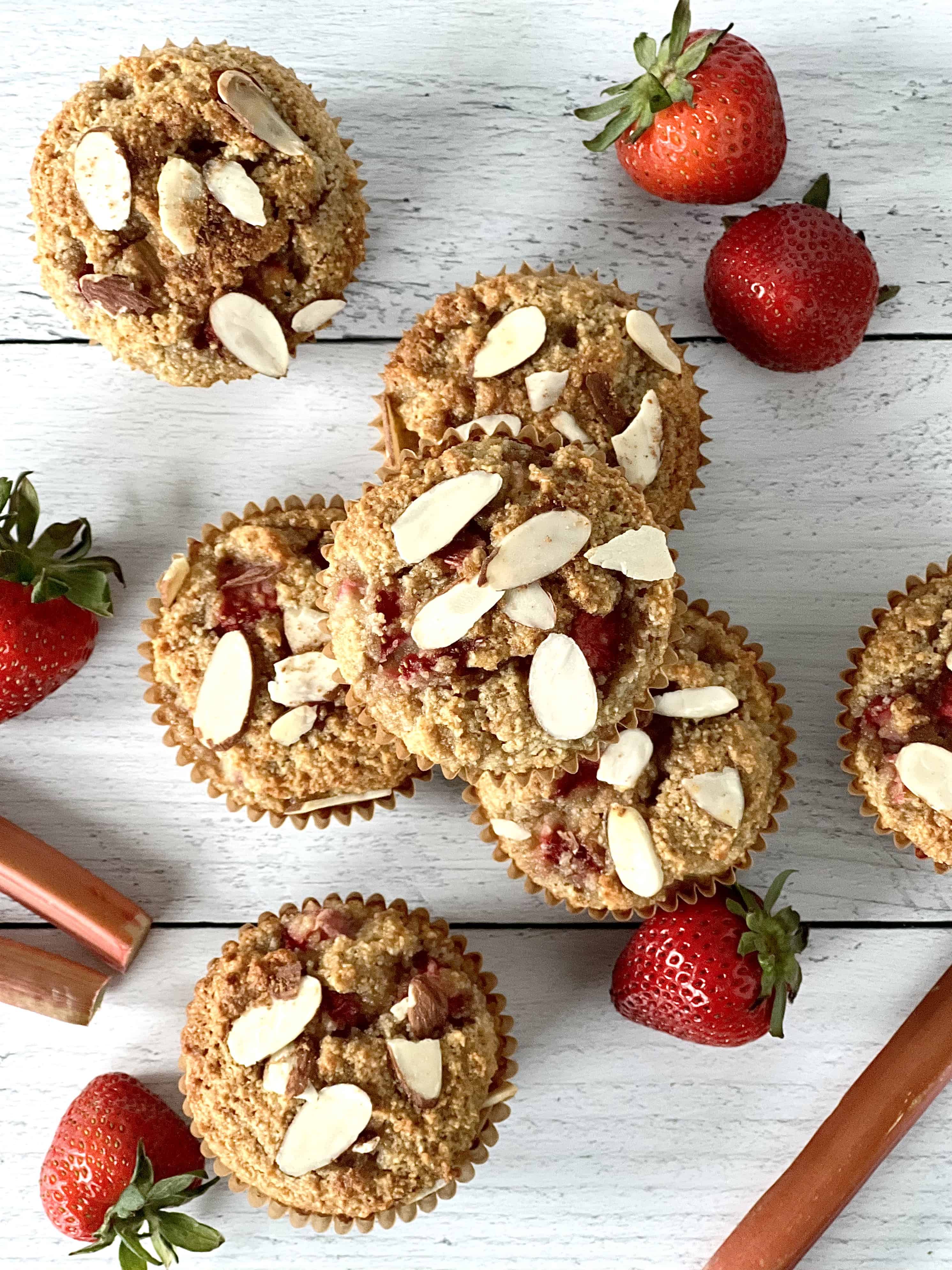 grain-free muffins with strawberries and rhubarb, plus sliced almonds on top, on a white wooden table with rhubarb stalks and strawberries