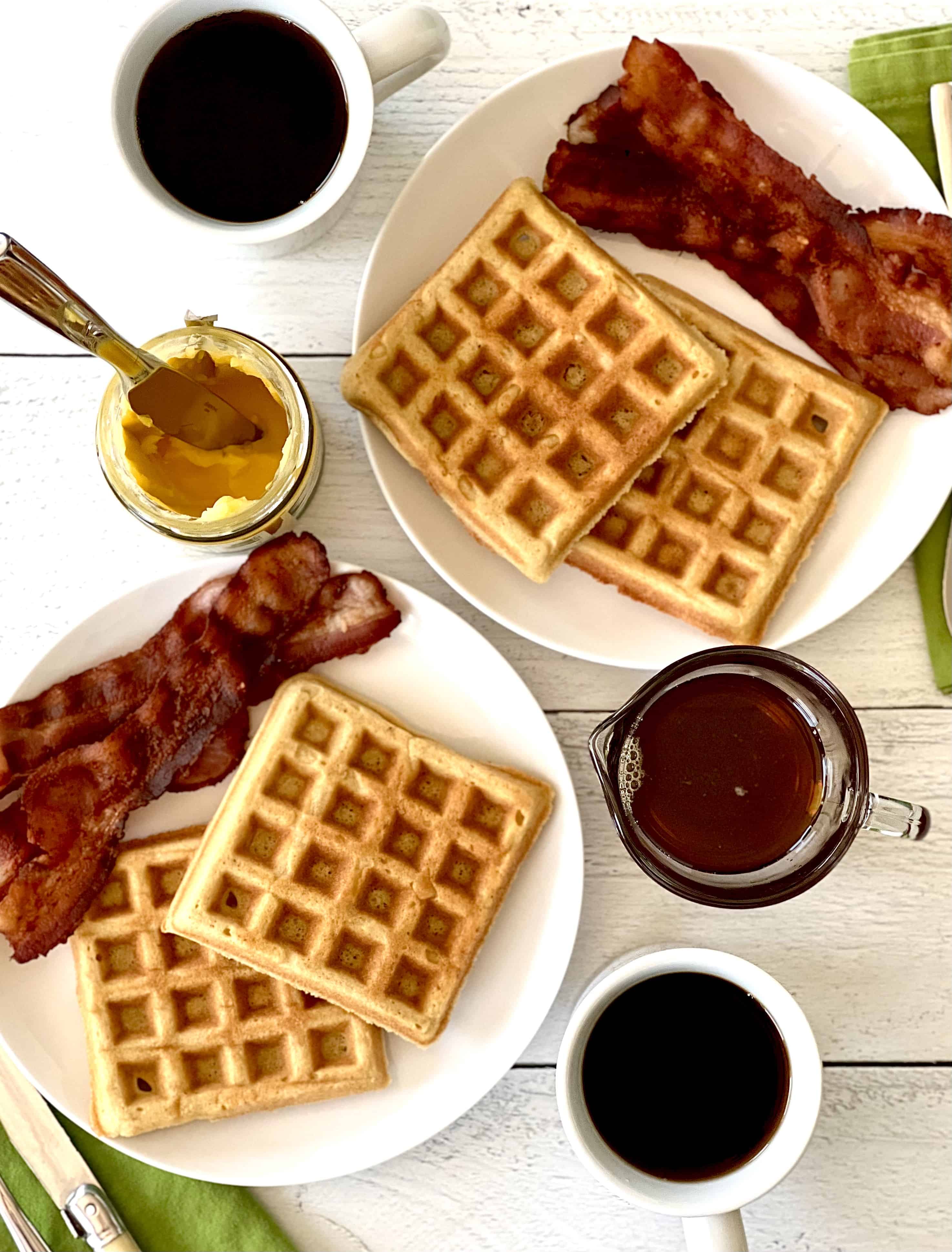 gluten-free waffles on white plates with bacon, next to white mugs of coffee, a glass pitcher of maple syrup and a jar of ghee
