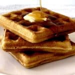 healthy waffles stacked on a white plate, topped with a pat of butter and with maple syrup getting poured on top