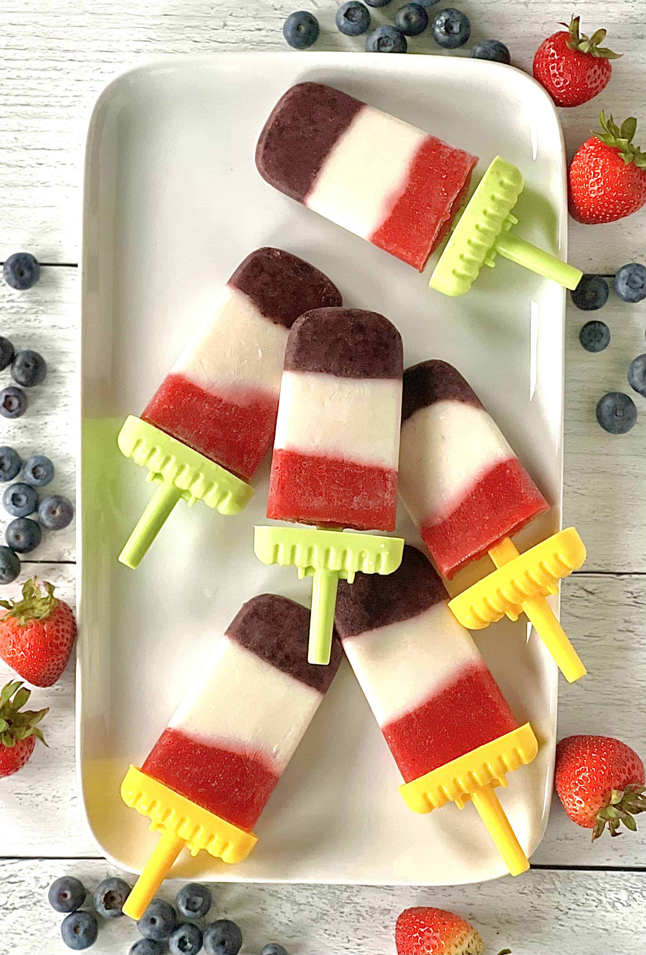 healthy homemade popsicles with layers of blueberries, strawberries and yogurt on a white platter surrounded by blueberries and strawberries