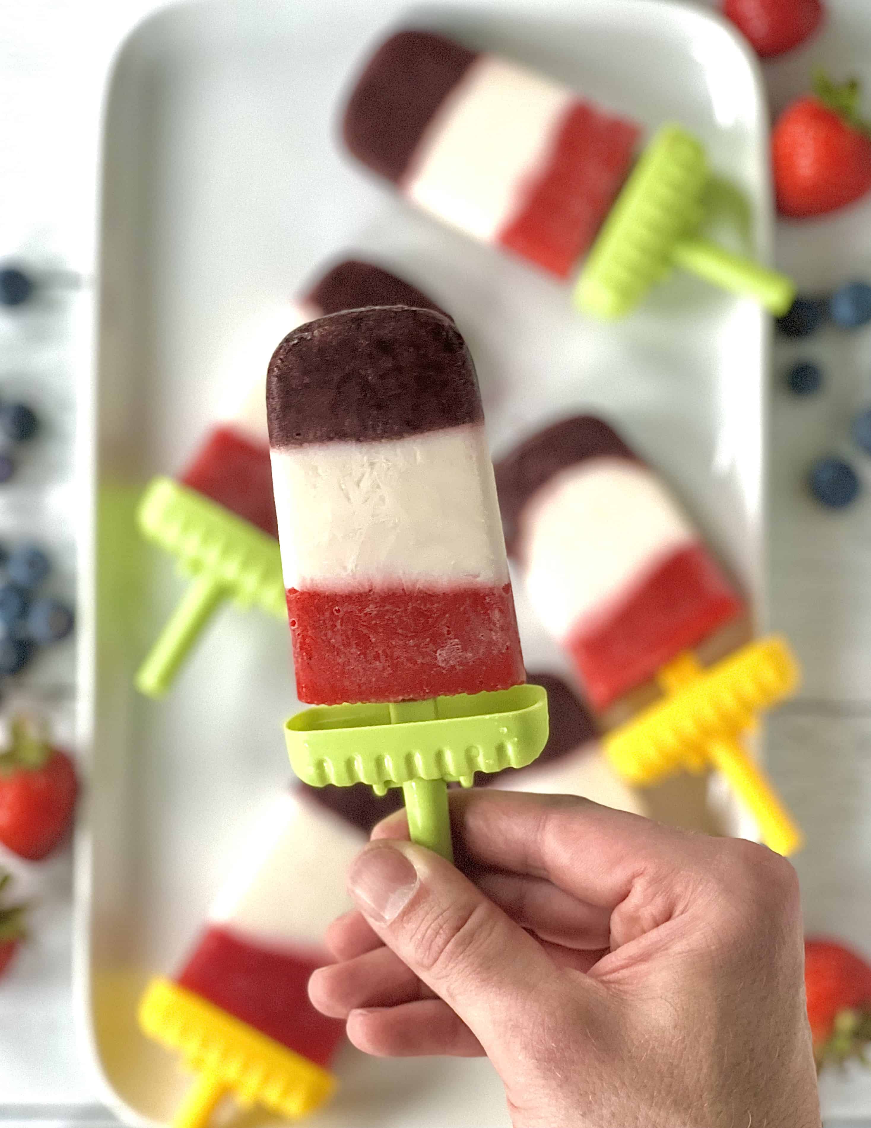 a hand holding up a real fruit popsicle with layers of blueberries, strawberries and yogurt above a white platter of more popsicles