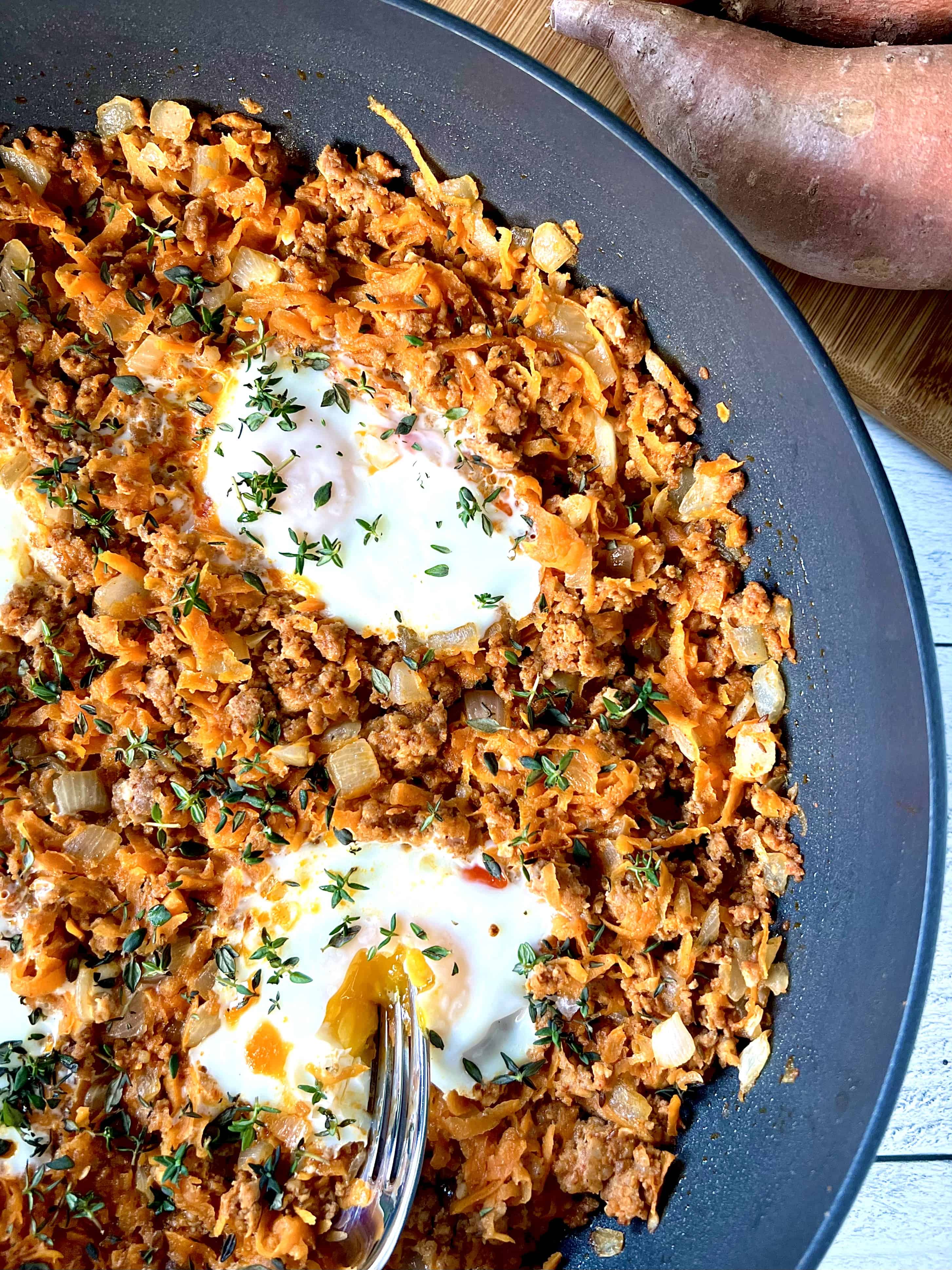 Whole30 sweet potato hash in a large skillet with 2 eggs and a fork cutting into one of the eggs