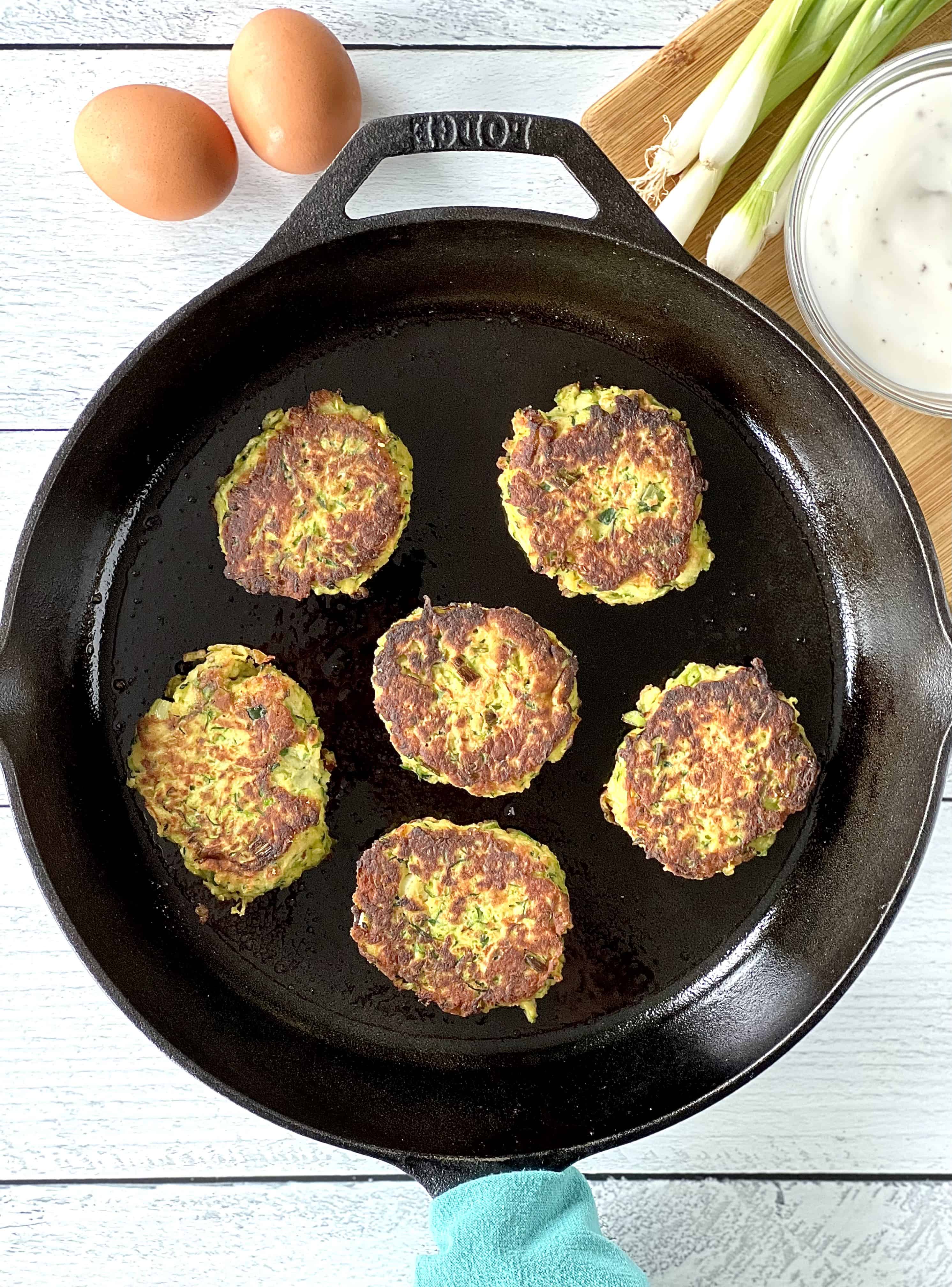 healthy zucchini fritters in a cast iron skillet with a blue towel around the handle, sitting on a white wooden table next to a bowl of yogurt sauce, scallions and eggs