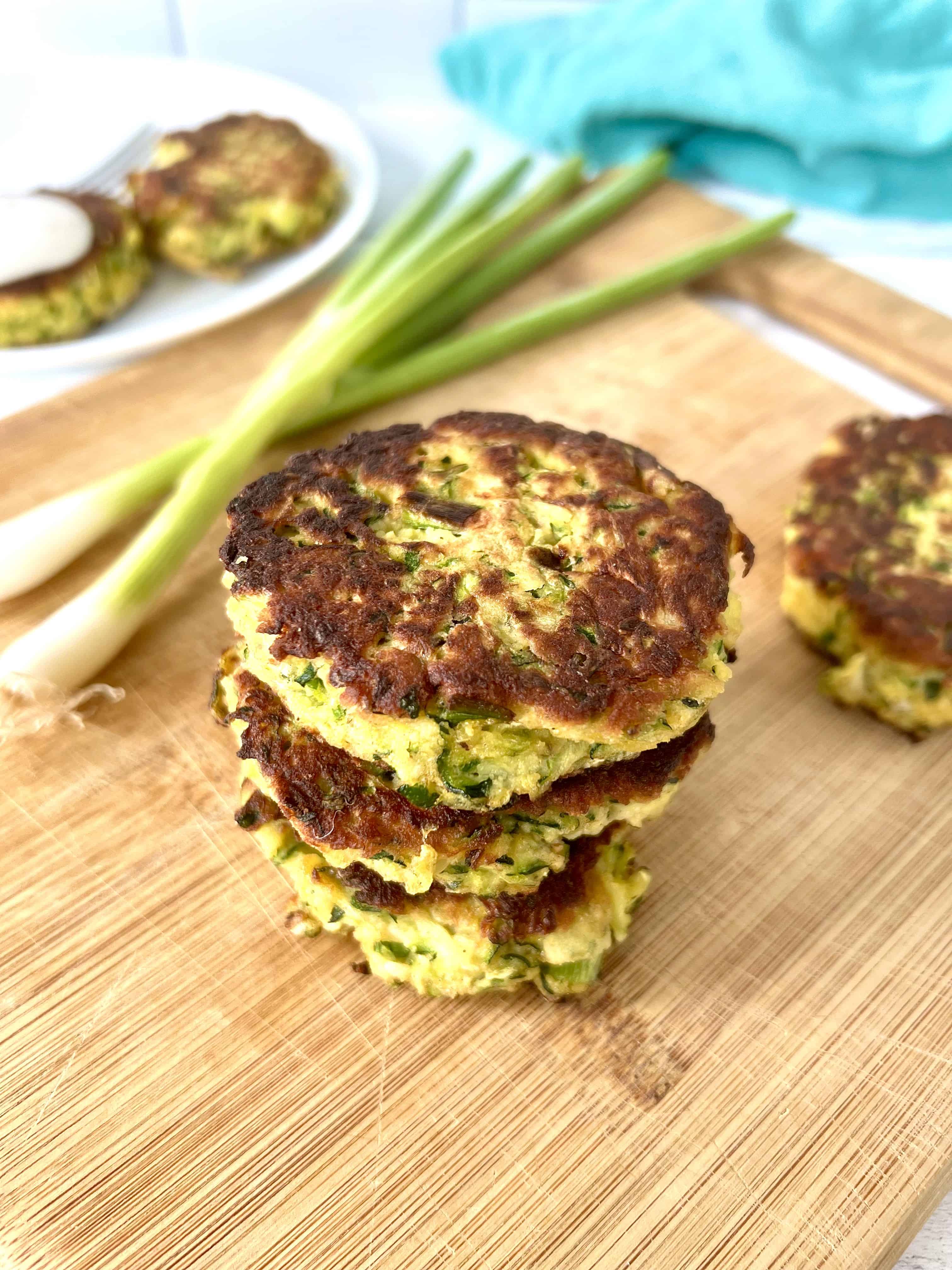 coconut flour zucchini fritters stacked on a wooden cutting board next to scallions and another fritter
