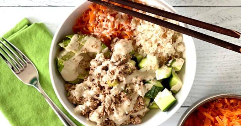 grain-free sushi bowl with canned tuna, vegetables, avocado and a creamy sauce with chopsticks on the rim of the bowl and a fork on a green napkin next to the white bowl