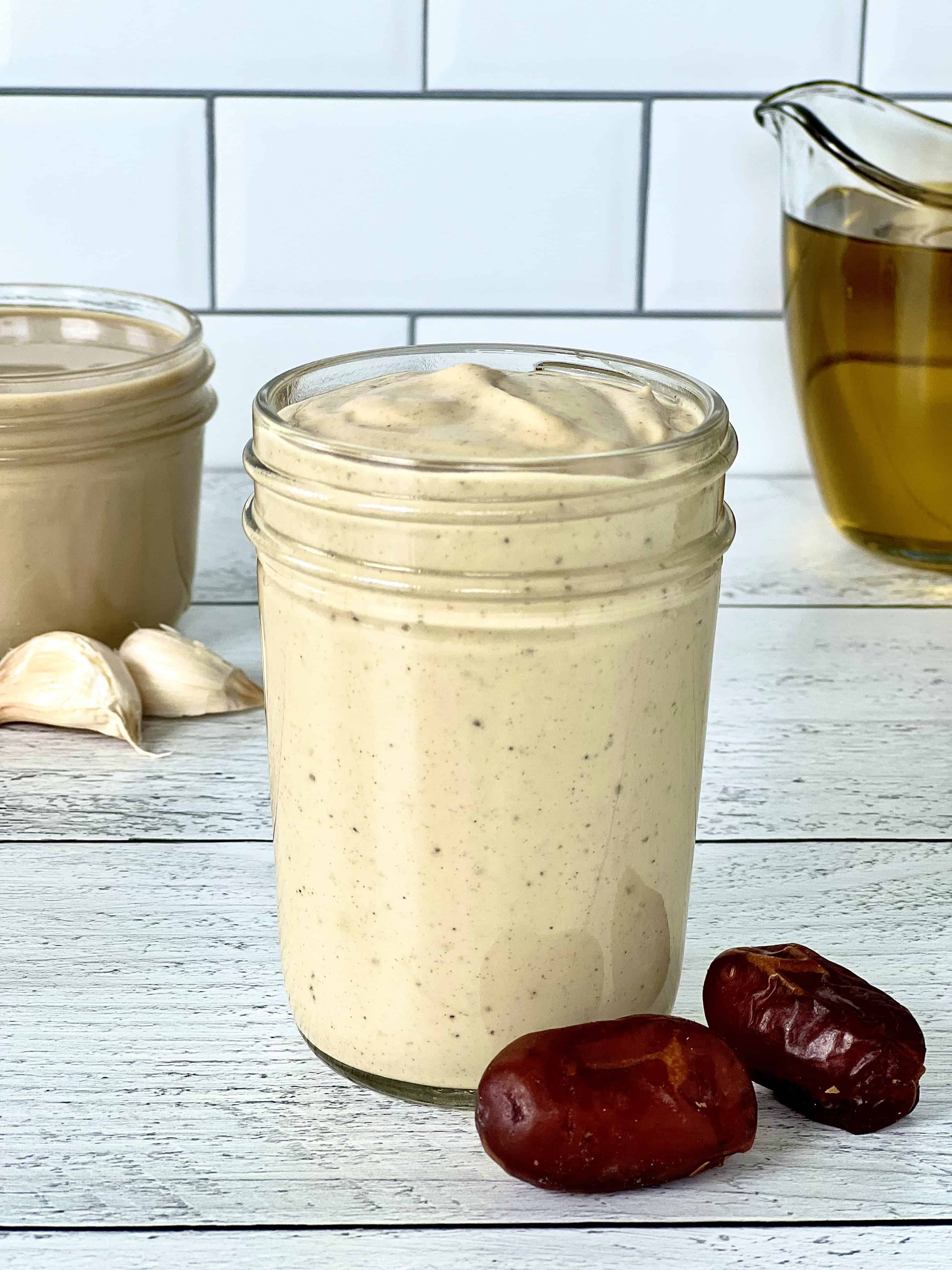 Whole30 tahini dressing in a mason jar next to some dates, unpeeled garlic cloves, a mason jar of tahini and a glass pitcher of olive oil