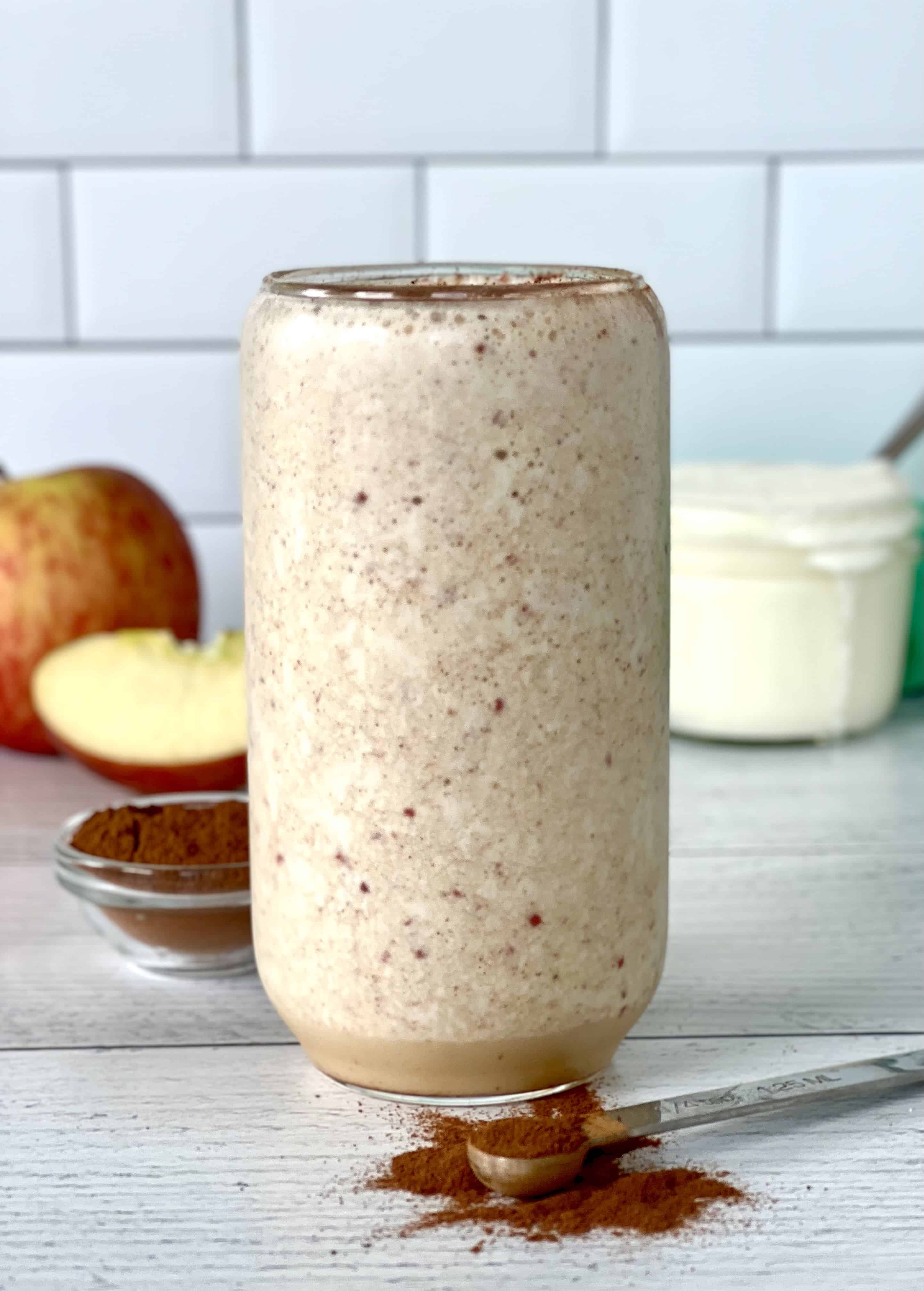 dairy-free apple yogurt smoothie in a tall glass topped with cinnamon, behind a measuring spoon spilling cinnamon on the table