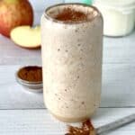 a vegan apple smoothie in a tall glass topped with cinnamon, next to a measuring spoon full of cinnamon spilling some on the table