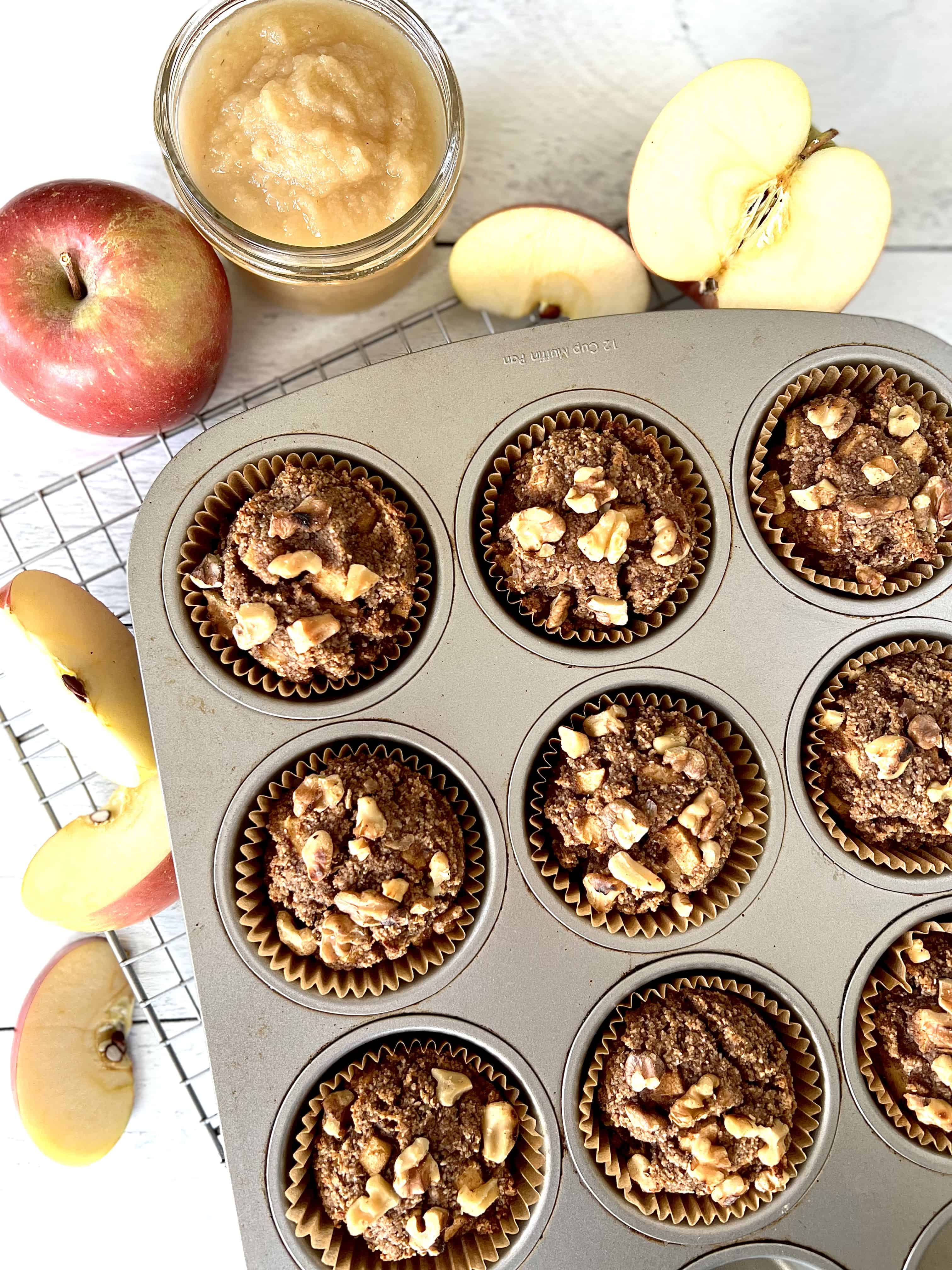 gluten-free apple muffins made with almond flour sitting in a metal muffin pan on a cooling rack surrounded by apple slices, an apple and a jar of applesauce