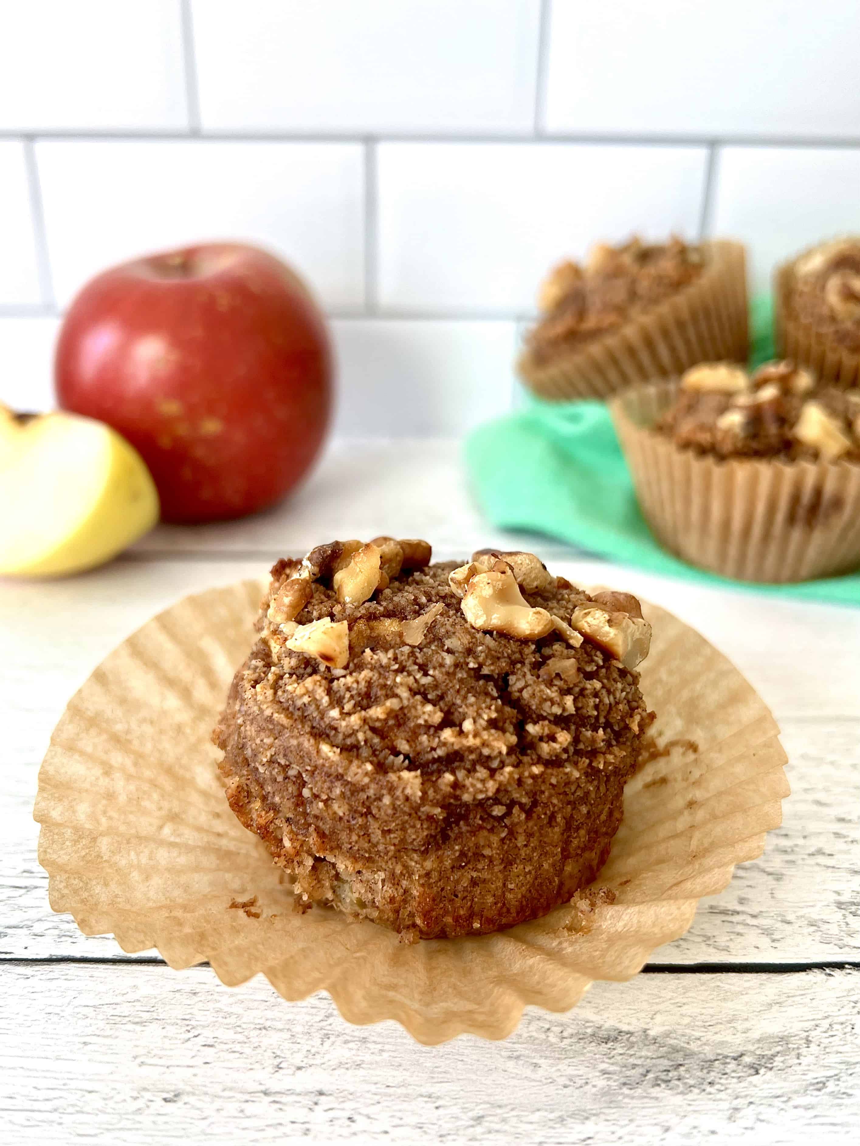 An Paleo muffin with apple chunks and walnuts on top on a white wooden table with its muffin liner pulled away