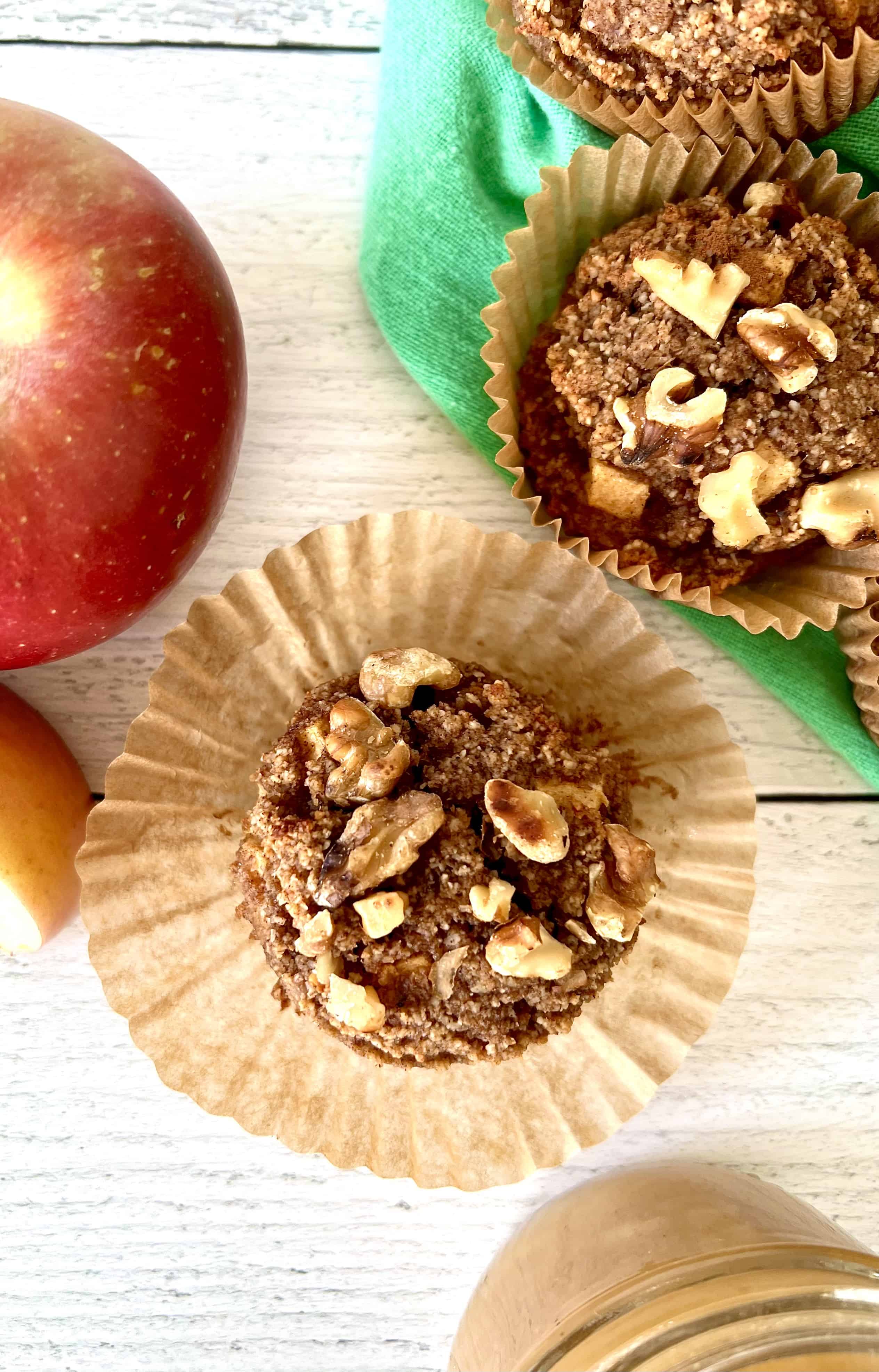 a grain-free apple muffin with its liner pulled away as it sits on a white wooden table next to an apple and more muffins