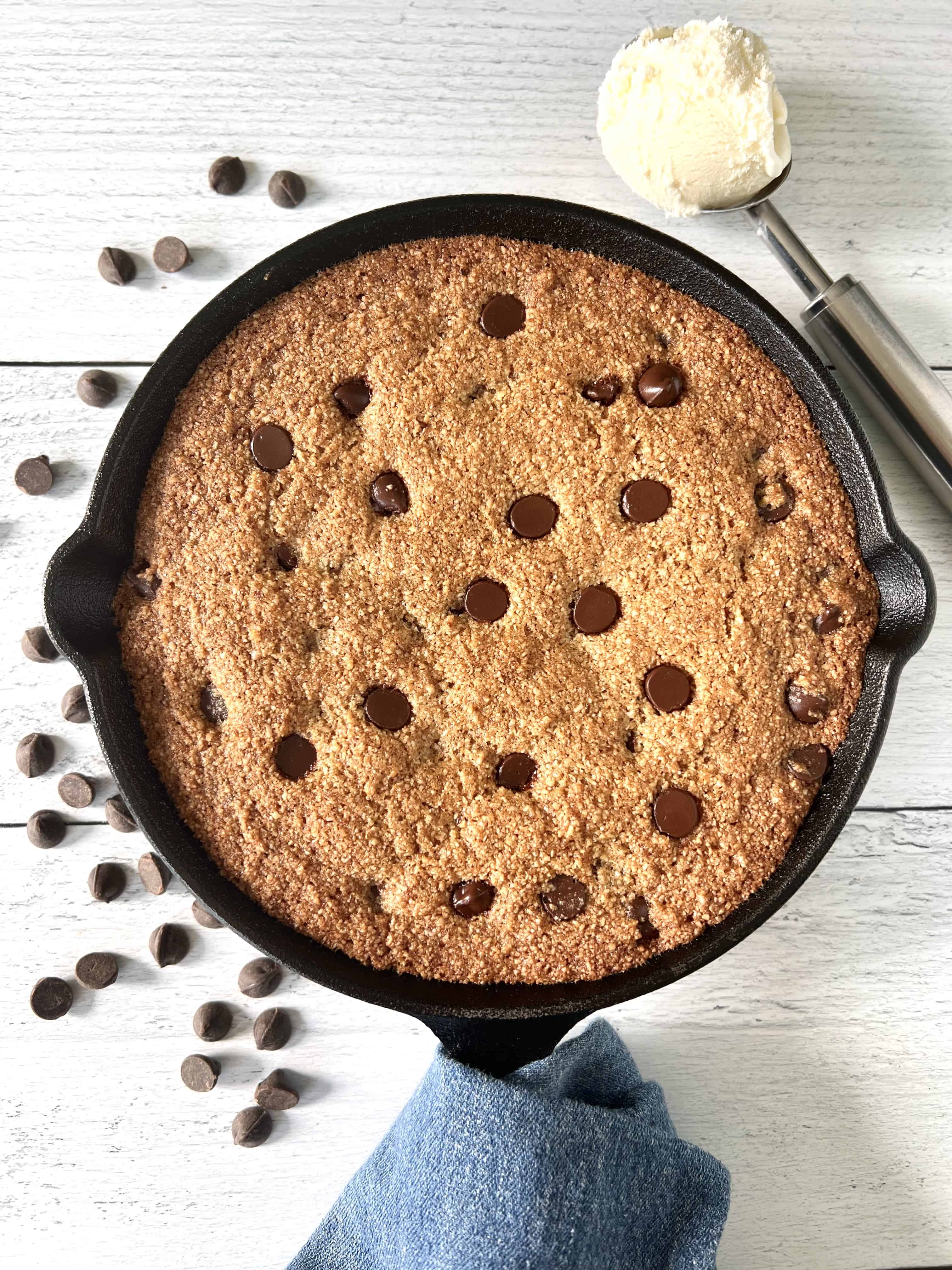 Gluten-free skillet cookie in a cast iron pan on a white wooden table strewn with chocolate chips and an ice cream scoop holding vanilla ice cream