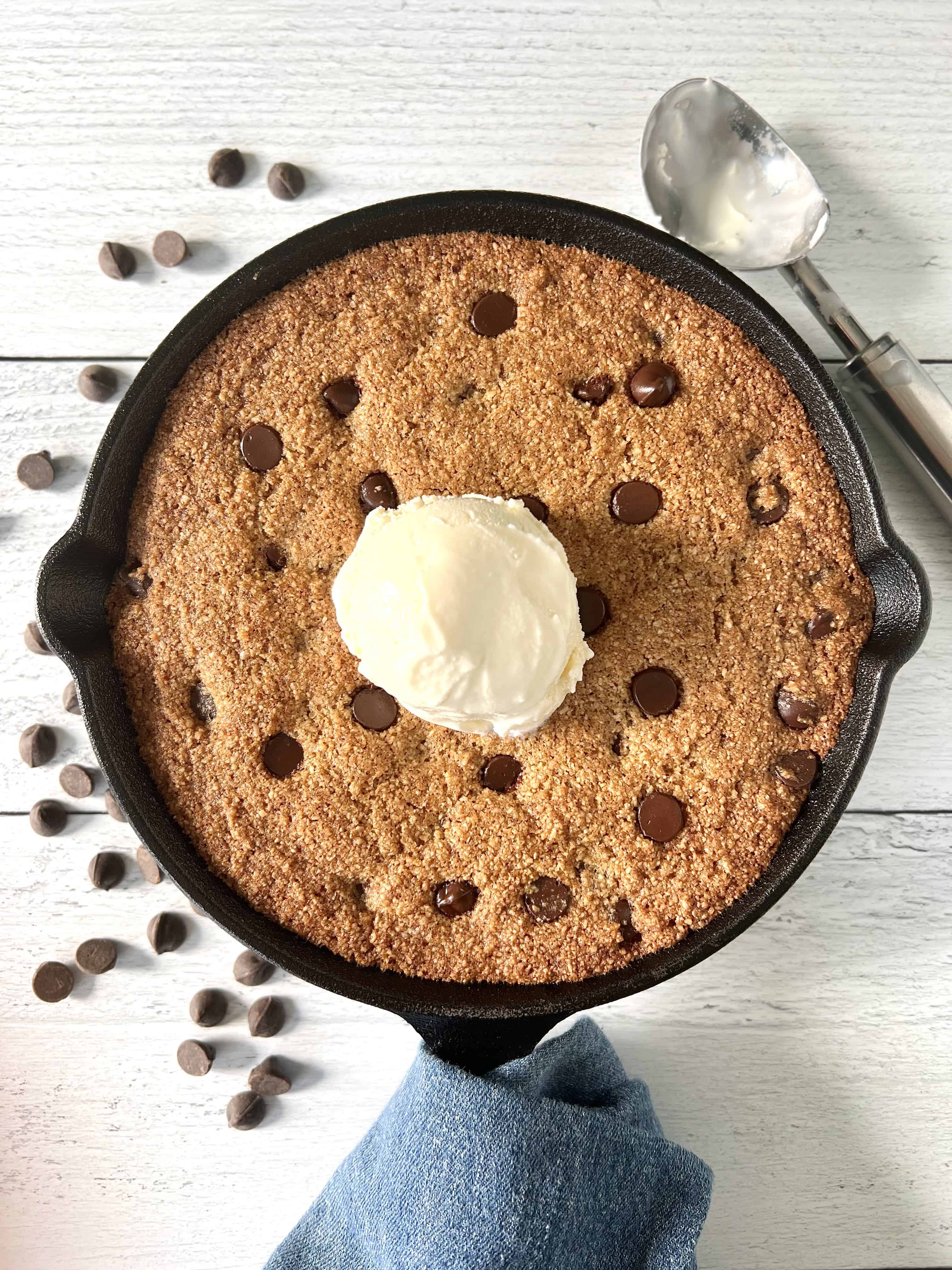 almond flour skillet cookie in a cast iron pan, topped with a scoop of vanilla ice cream, on a white wooden table strewn with chocolate chips and an ice cream scoop