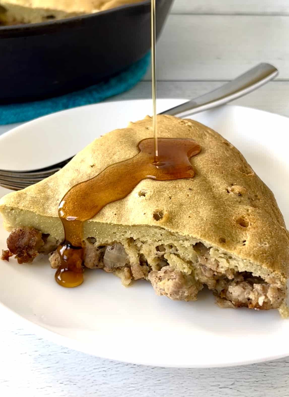 A slice of gluten-free pancake and sausage pie on a white plate getting drizzled with maple syrup