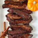 candied bacon laid out on a white rectangular platter with a mini pumpkin and cinnamon sticks