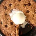 Grain-Free Skillet Cookie in a cast iron pan with a slice missing and a scoop of vanilla ice cream melting on top