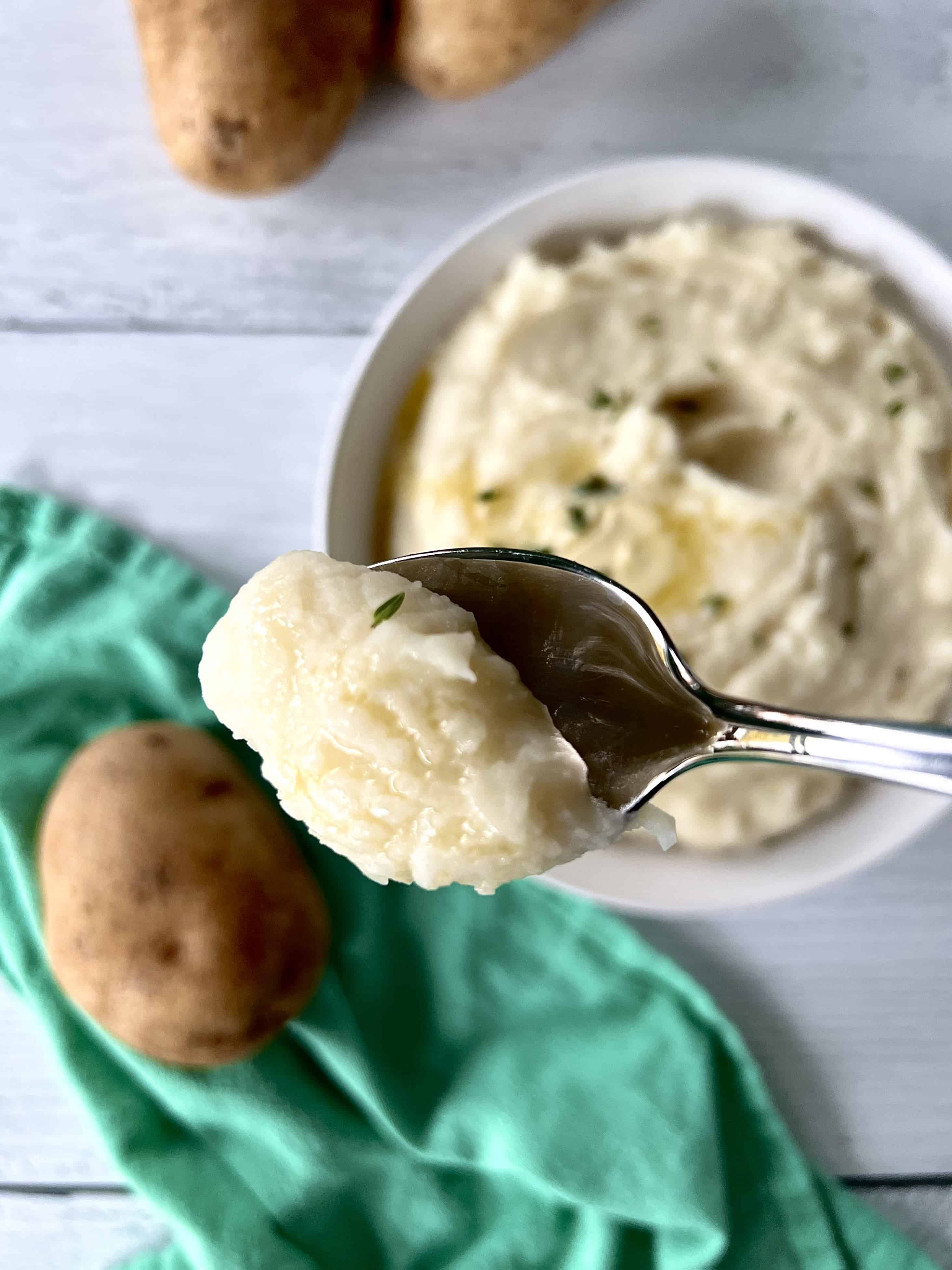 a spoon holding up creamy homemade mashed potatoes over a bowl of mashed potatoes, plus a green napkin with potatoes