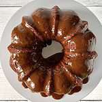 gluten-free pumpkin bundt cake coated in a dripping caramel sauce, on a white cake stand over a white wooden table