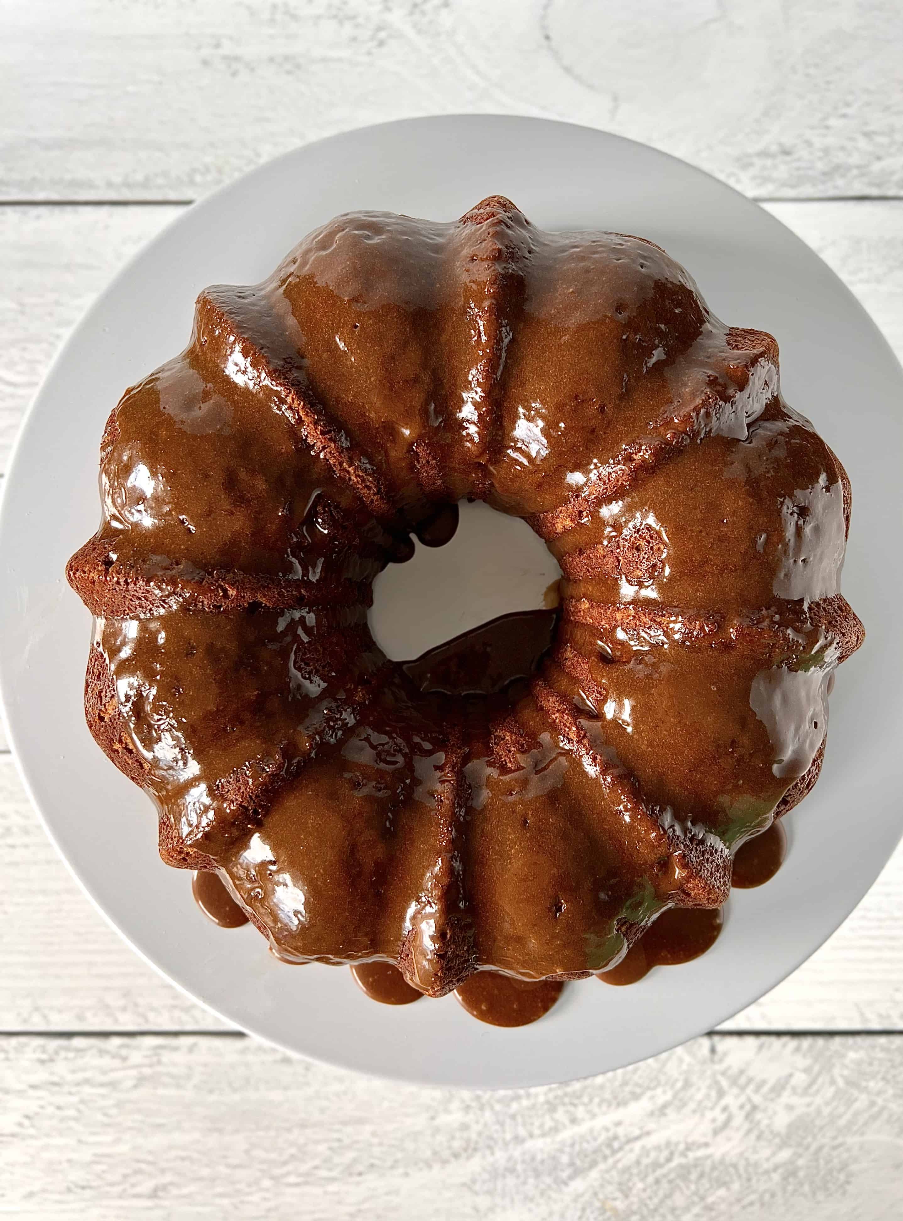 gluten-free pumpkin bundt cake coated in a dripping caramel sauce, on a white cake stand over a white wooden table