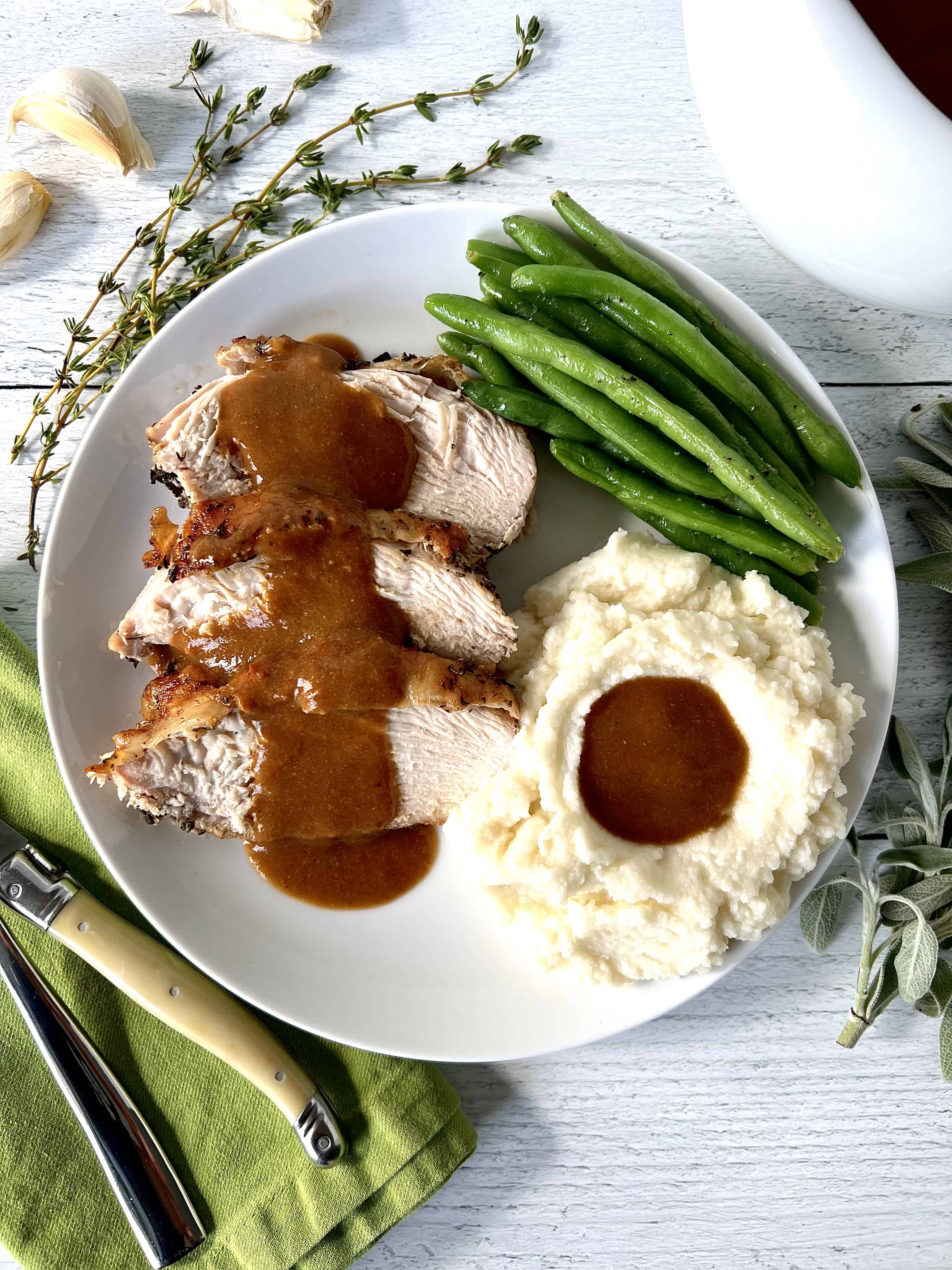 keto turkey breast slices on a white plate with gravy, mashed cauliflower and green beans, next to a green napkin with a fork and knife