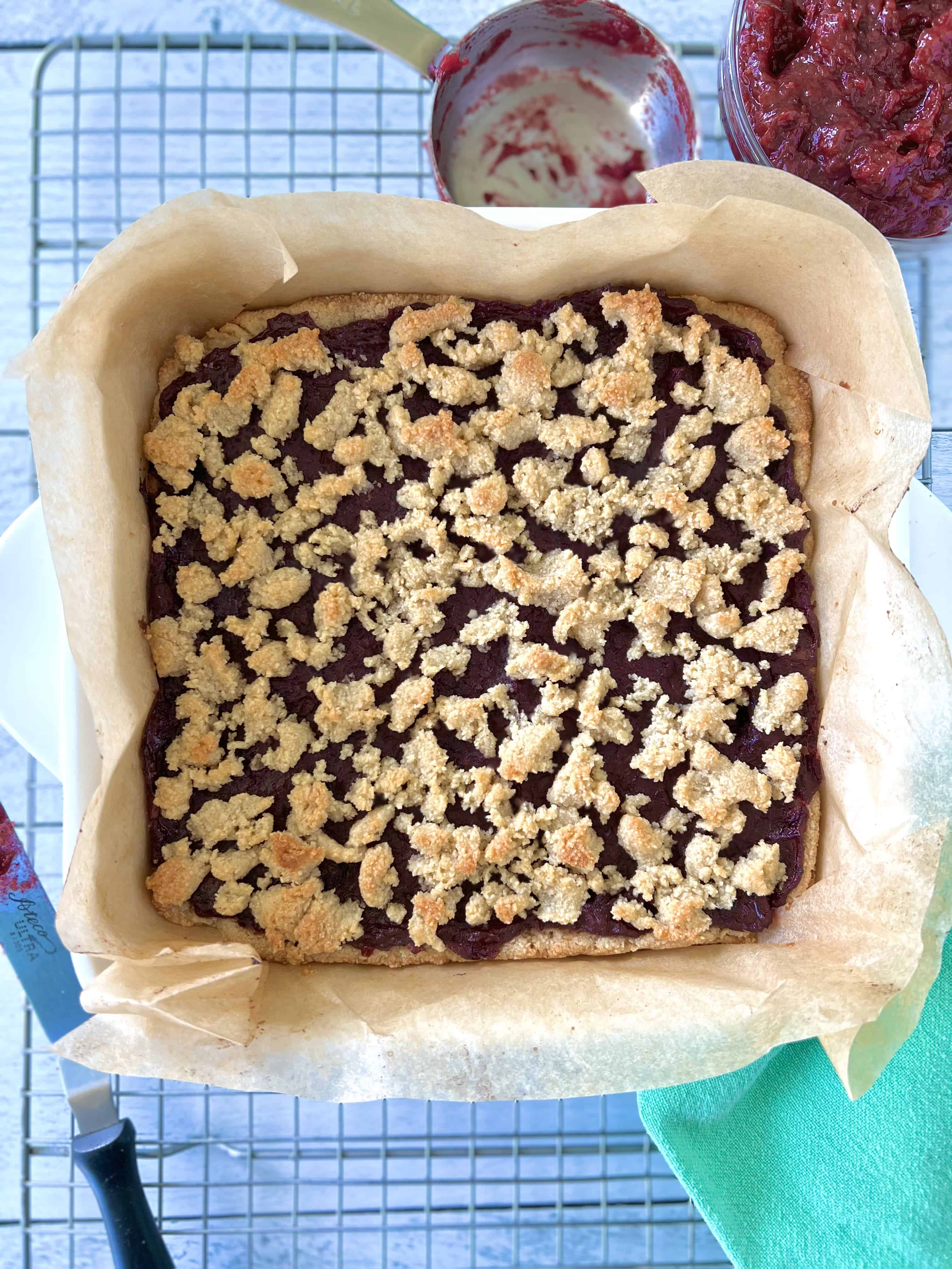 almond flour cranberry bars in a square baking dish lined with parchment paper, on a cooling rack with a small offset spatula, a green napkin, a bowl of cranberry sauce and a measuring cup