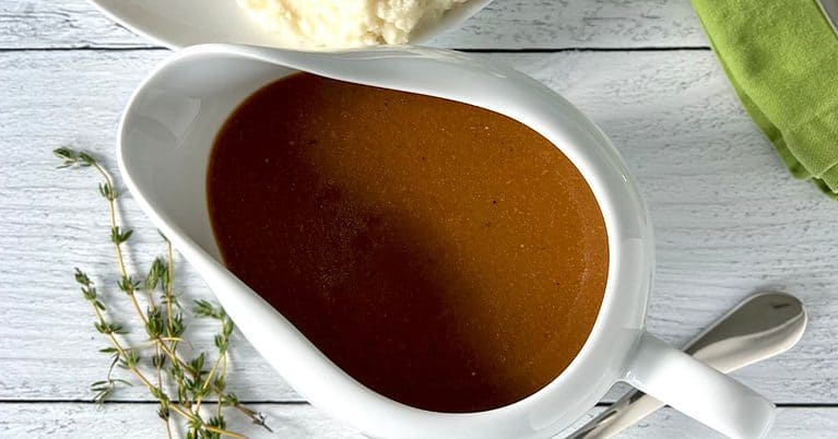 Whole30 gravy in a white gravy boat next to a silver lade, fresh thyme and a green napkin on a white wooden table