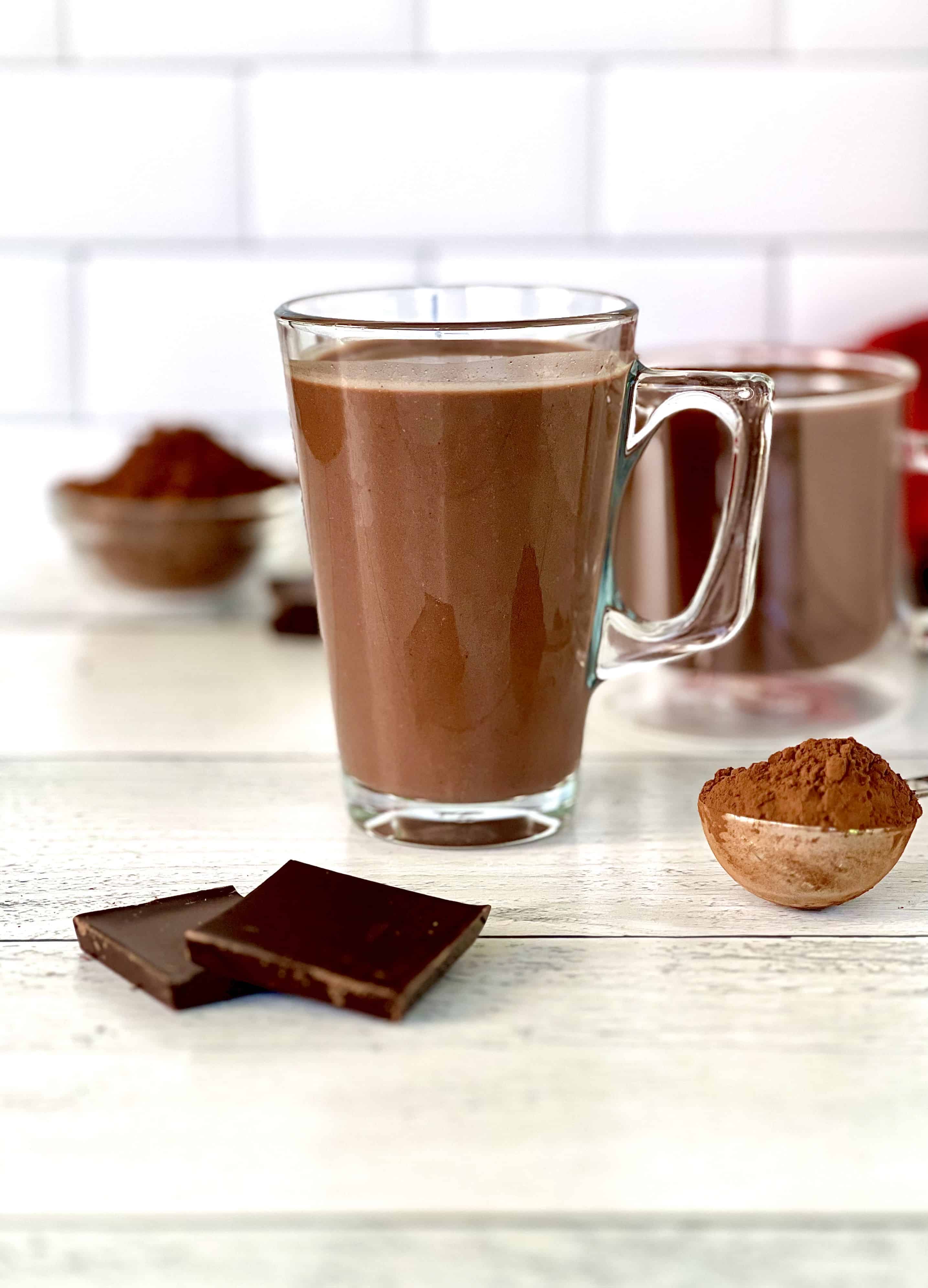 Hot cocoa in a glass mug with a glass mug of sipping chocolate in the background
