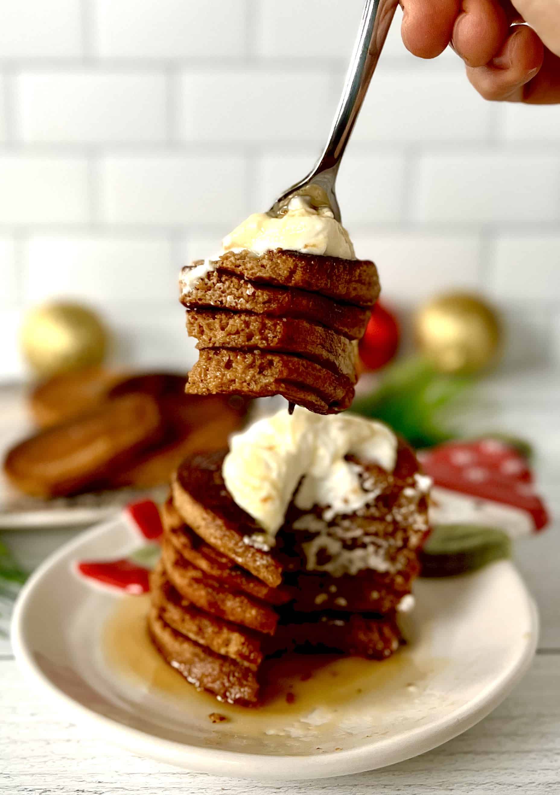 gluten-free gingerbread pancakes stacked on a snowman plate with yogurt and maple syrup on top, plus a fork holding up a bite of four layers and yogurt on top