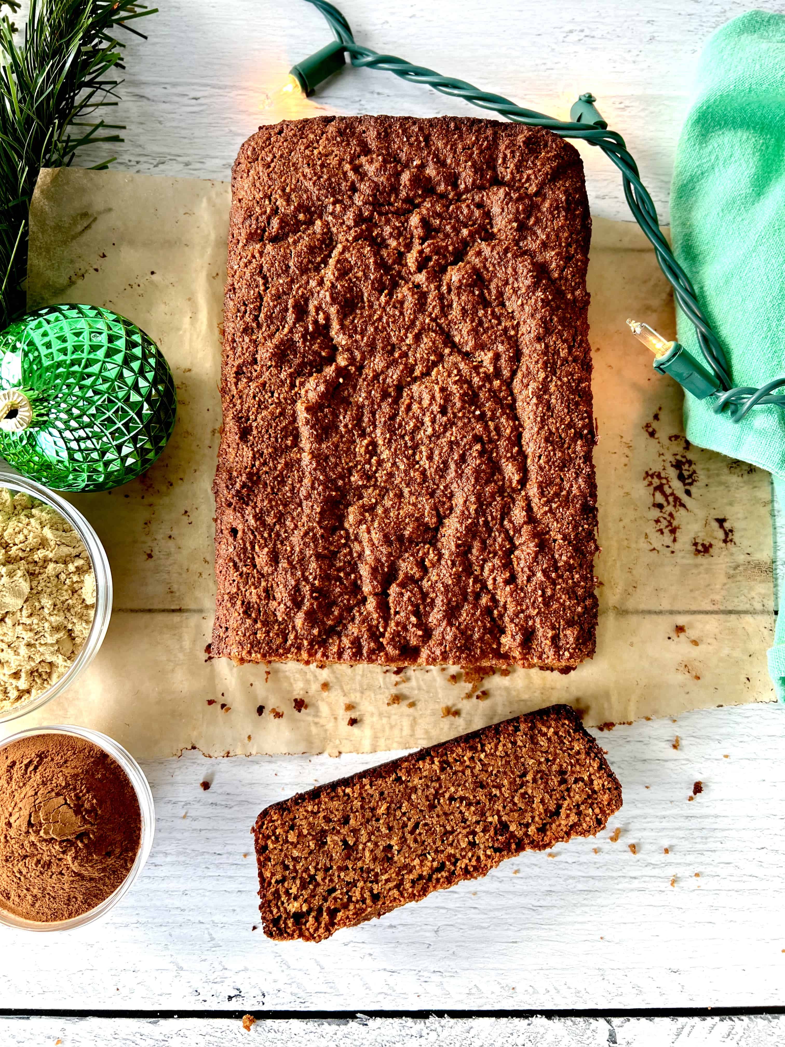 grain free gingerbread sliced on parchment paper on a white wooden table between Christmas lights, a green napkin, a green ornament and small bowls of spices