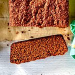 Almond flour gingerbread loaf sliced on a white wooden table between Christmas lights, a green napkin and small bowls of spices