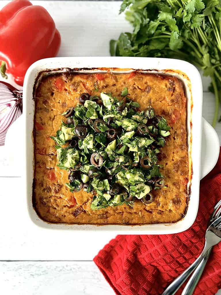 A gluten-free dairy-free casserole with chorizo sausage in a white baking dish next to a bunch of parsley, a red onion, a red bell pepper and a red towel with 2 forks on it.