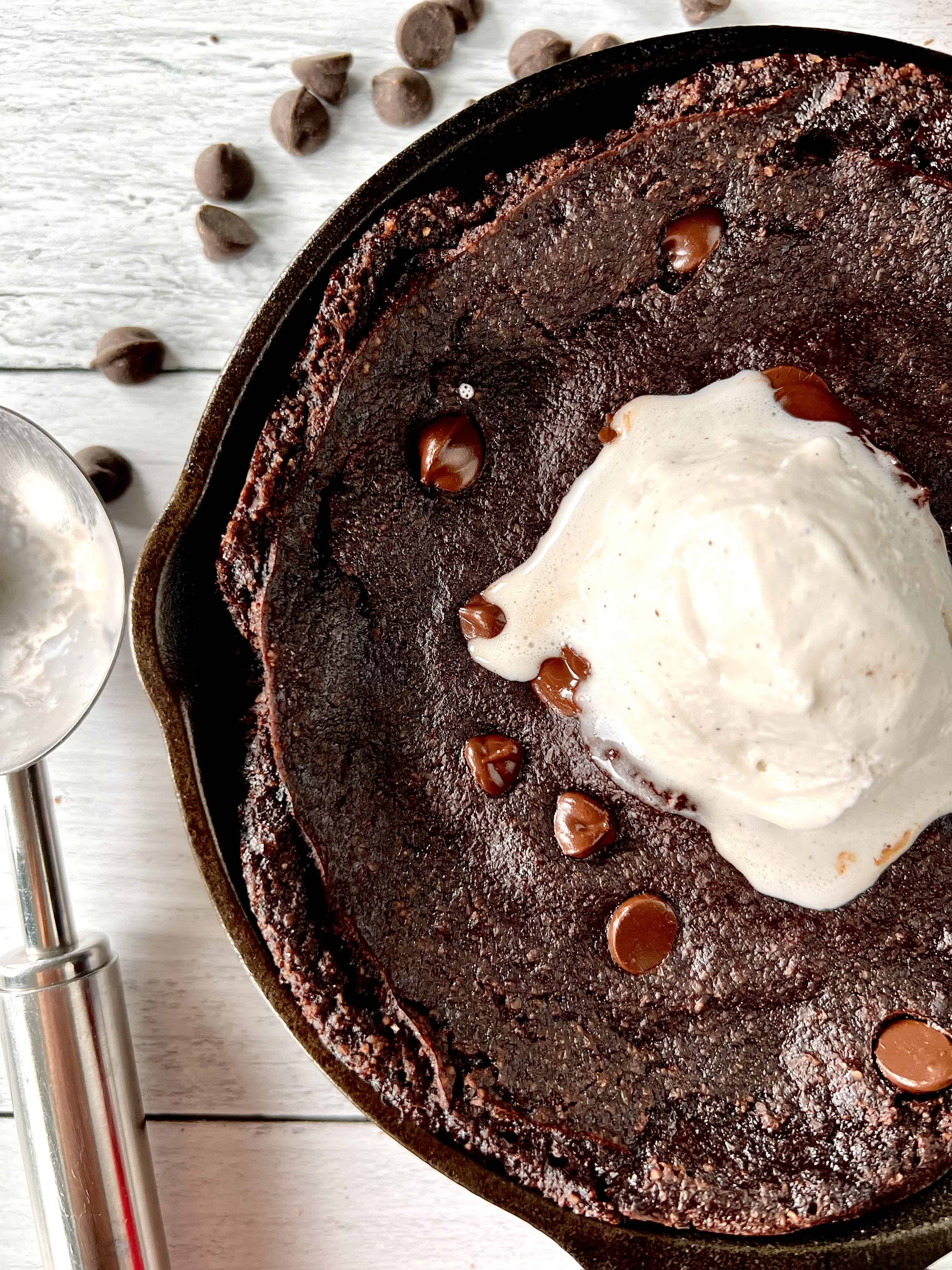 Flourless chocolate skillet cake topped with chocolate chips and vanilla ice cream, in a cast iron skillet next to an ice cream scoop.