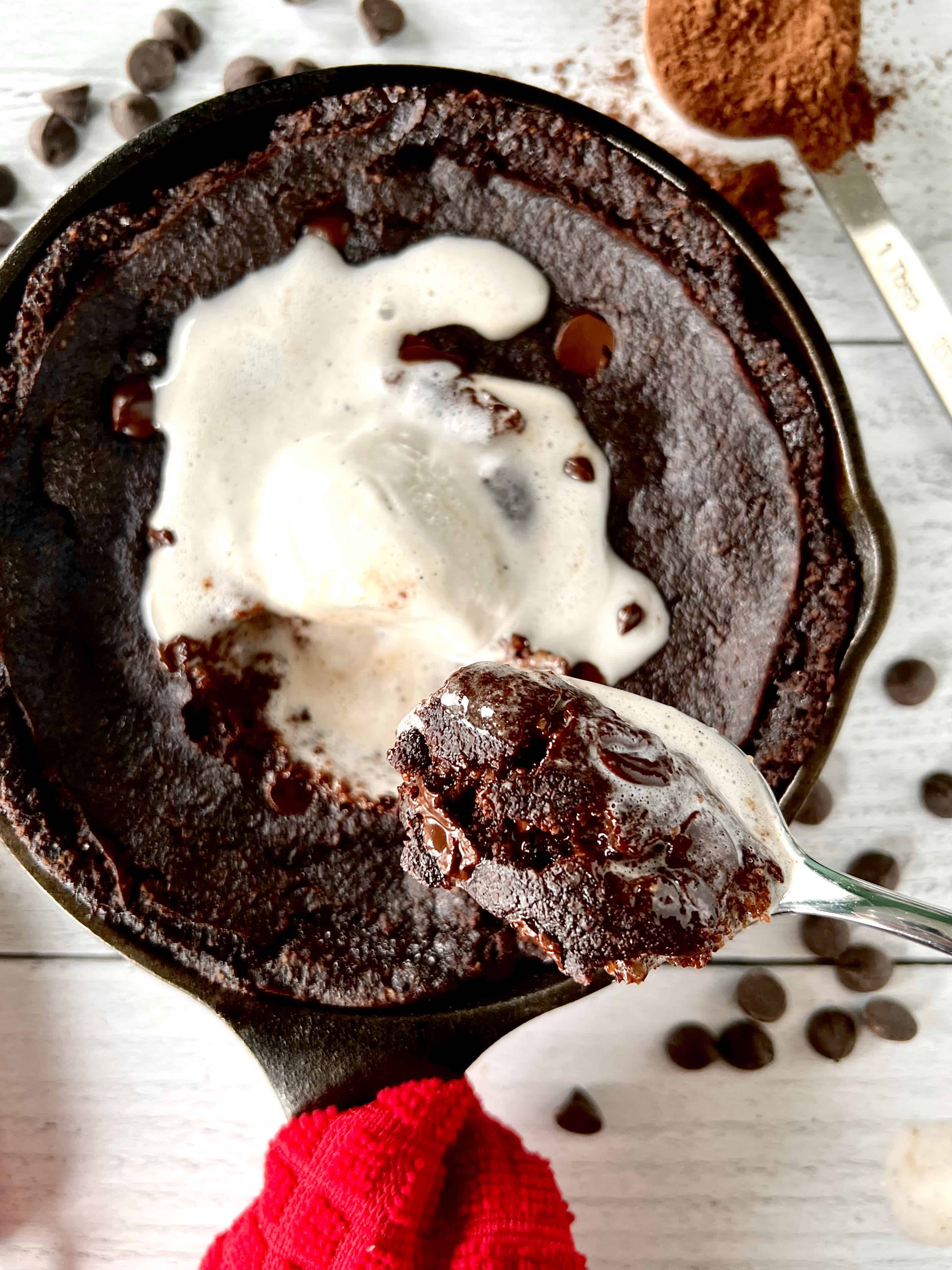 Healthy chocolate cake in a cast iron skillet topped with vanilla ice cream, with a spoon holding up a scoop of the cake and ice cream.