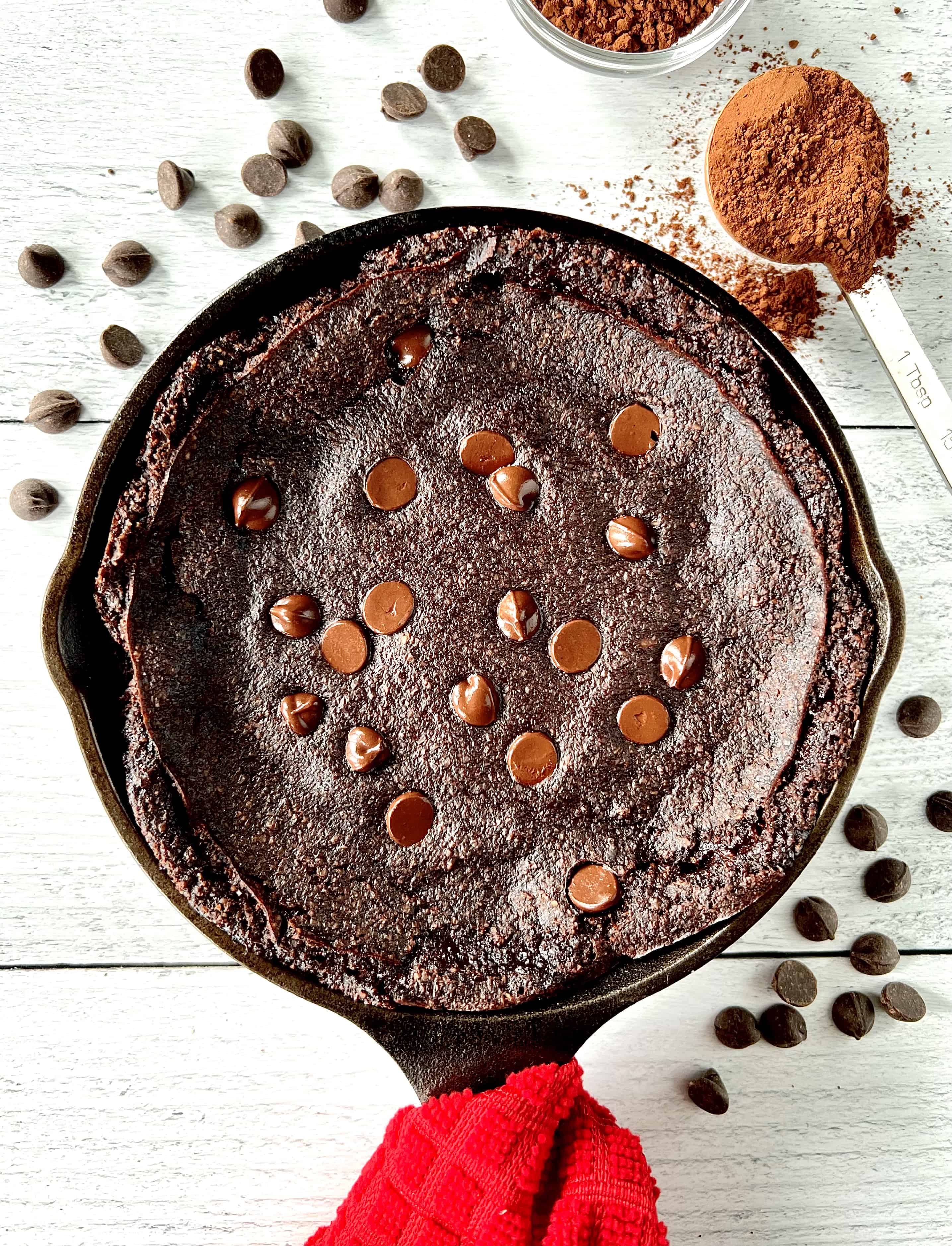 Chocolate vegan skillet cake topped with chocolate chips, in a cast iron skillet with a red towel around its handle, next to more chocolate chips and a bowl and spoon full of cocoa powder.