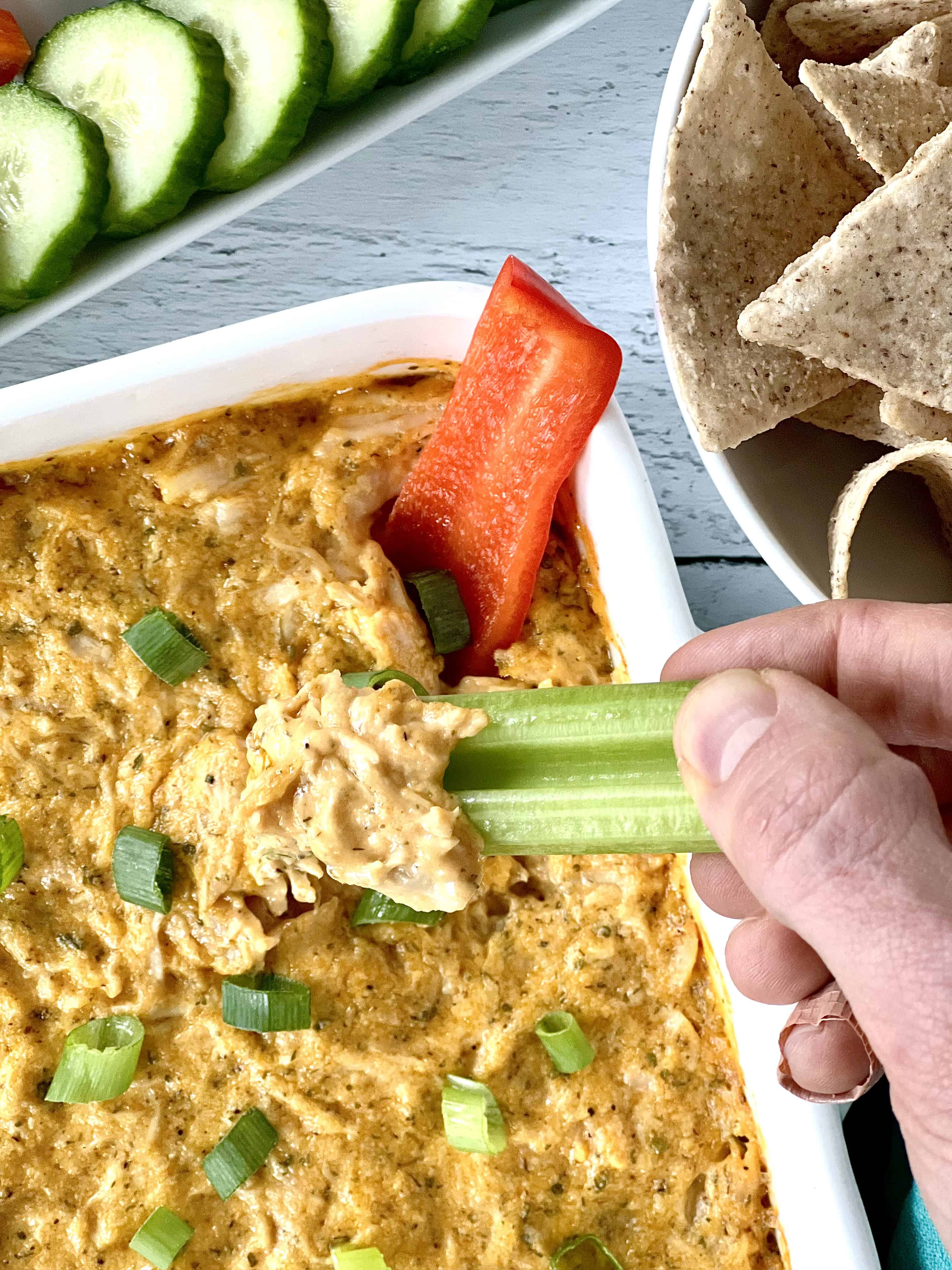 Healthier buffalo dip in a white square baking dish with a hand holding a celery stalk as it scoops up the dip