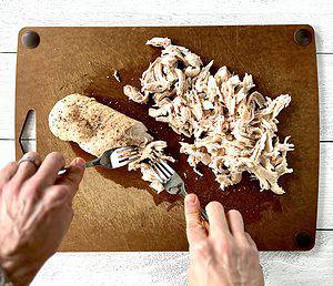 two hands holding forks as they shred two chicken breasts on a brown cutting board