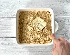Using a spatula to smooth Paleo buffalo chicken dip in a white square baking dish.