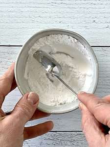 one hand holding a bowl of paleo baking powder while another hand mixes it with a measuring spoon