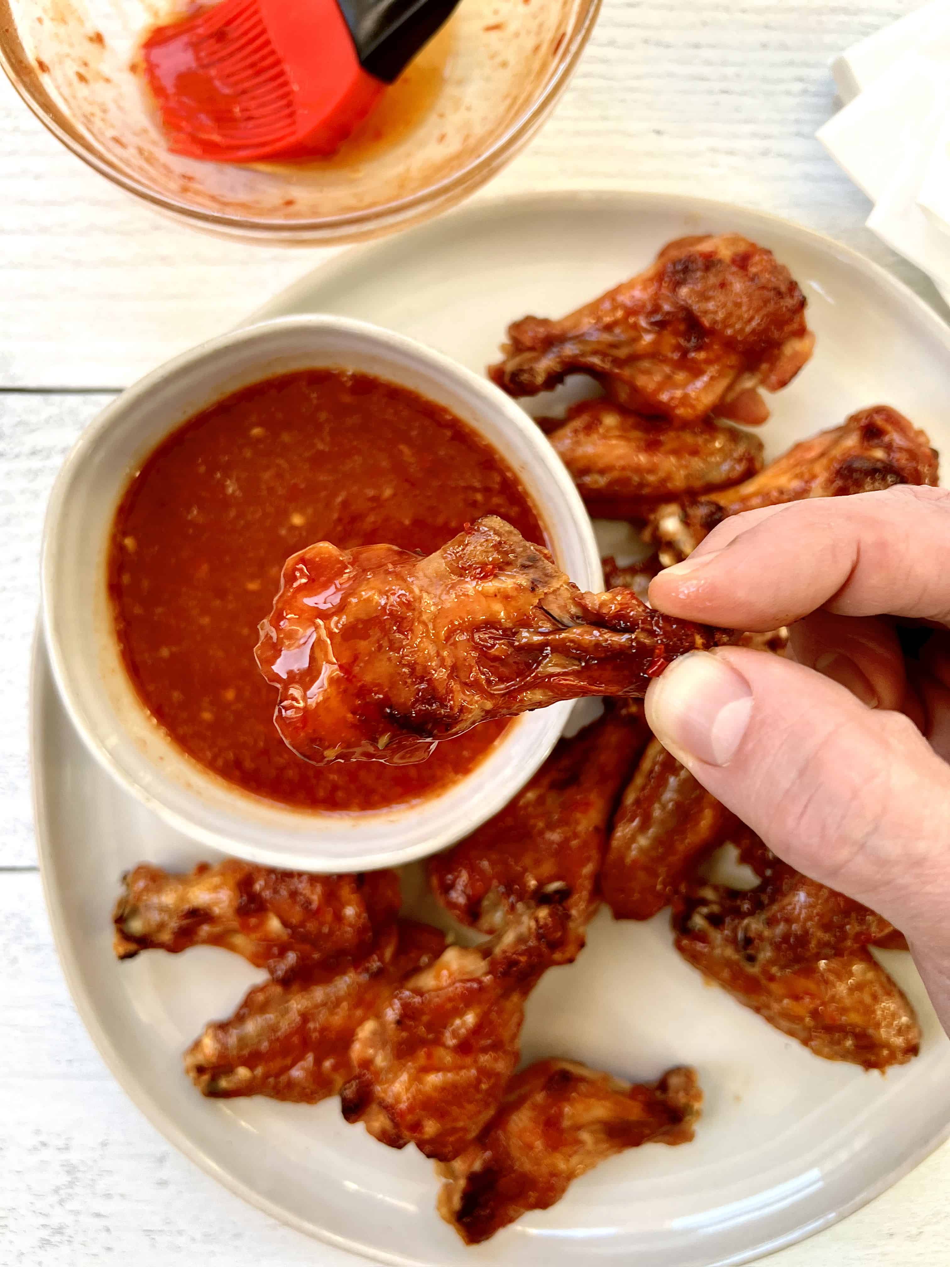 a hand holding up a hot wing above a platter of more hot wings and a small bowl of more hot chili sauce