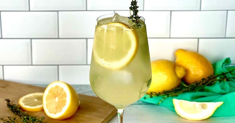 An Italian Prosecco cocktail in a wine glass with ice, a lemon slice and sprig of thyme.