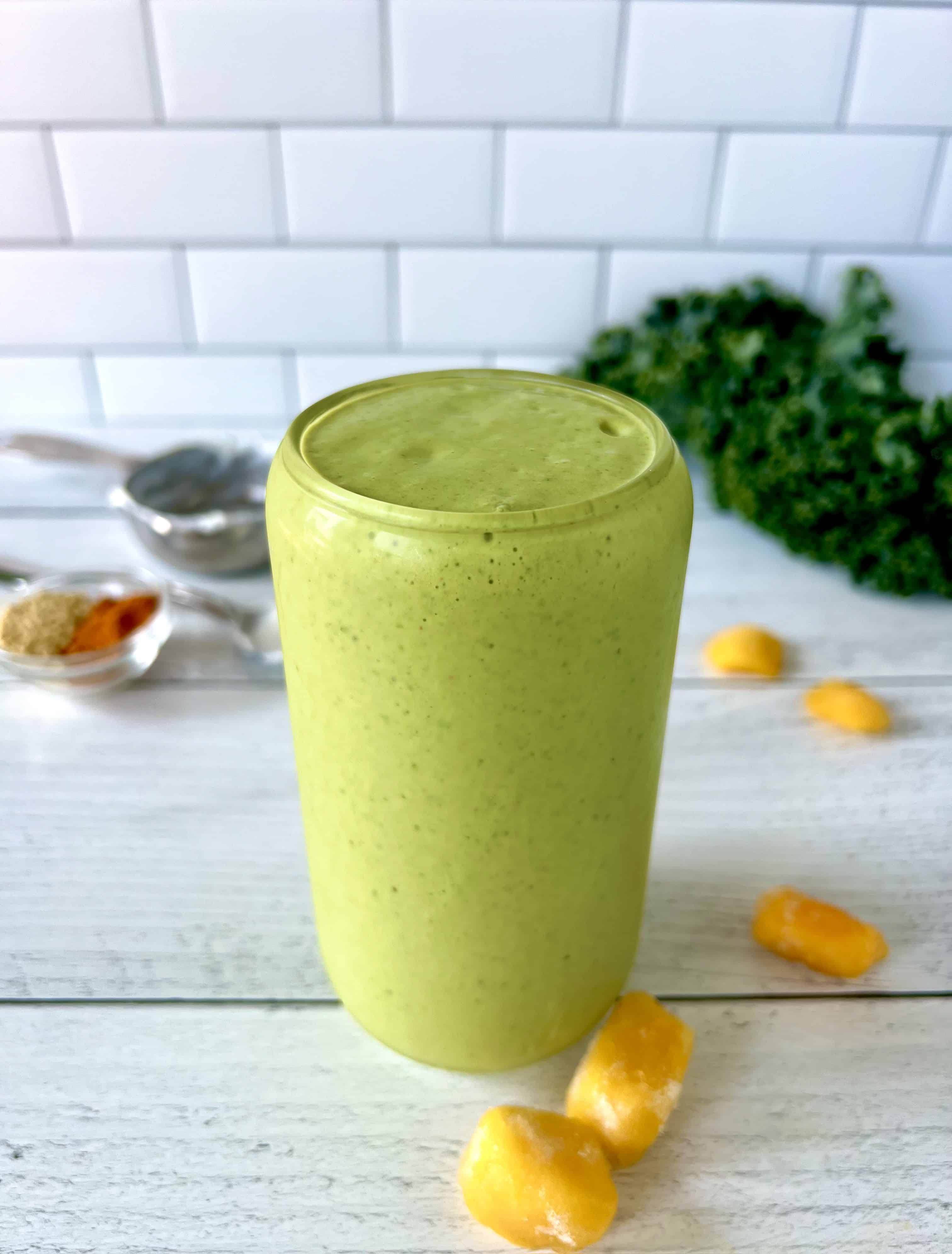 A Paleo green smoothie in a tall glass on a white wooden table next to frozen mango chunks and large kale leaves.