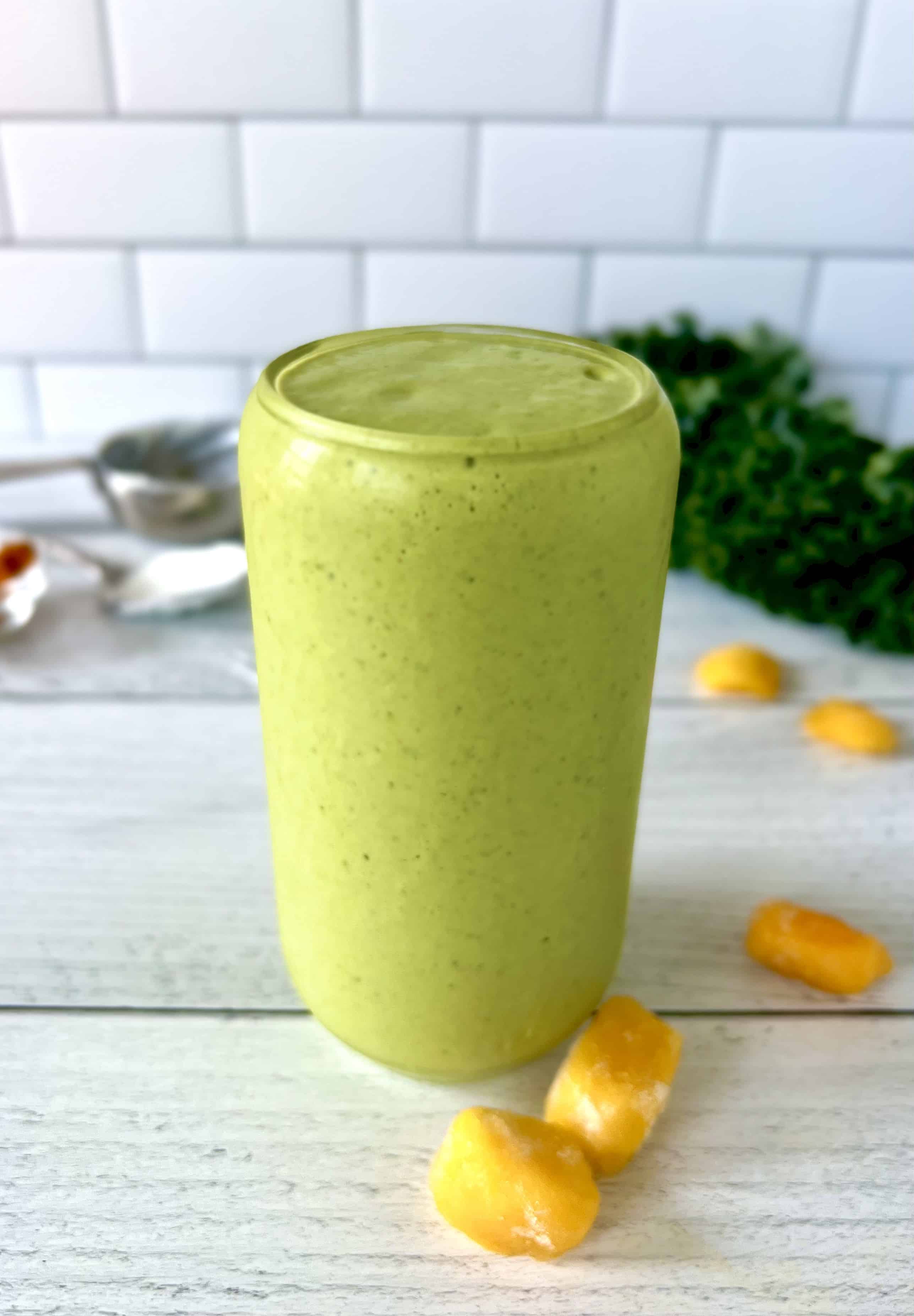A tall glass of a healthy kale smoothie on a white wooden table next to frozen mango chunks and large kale leaves.