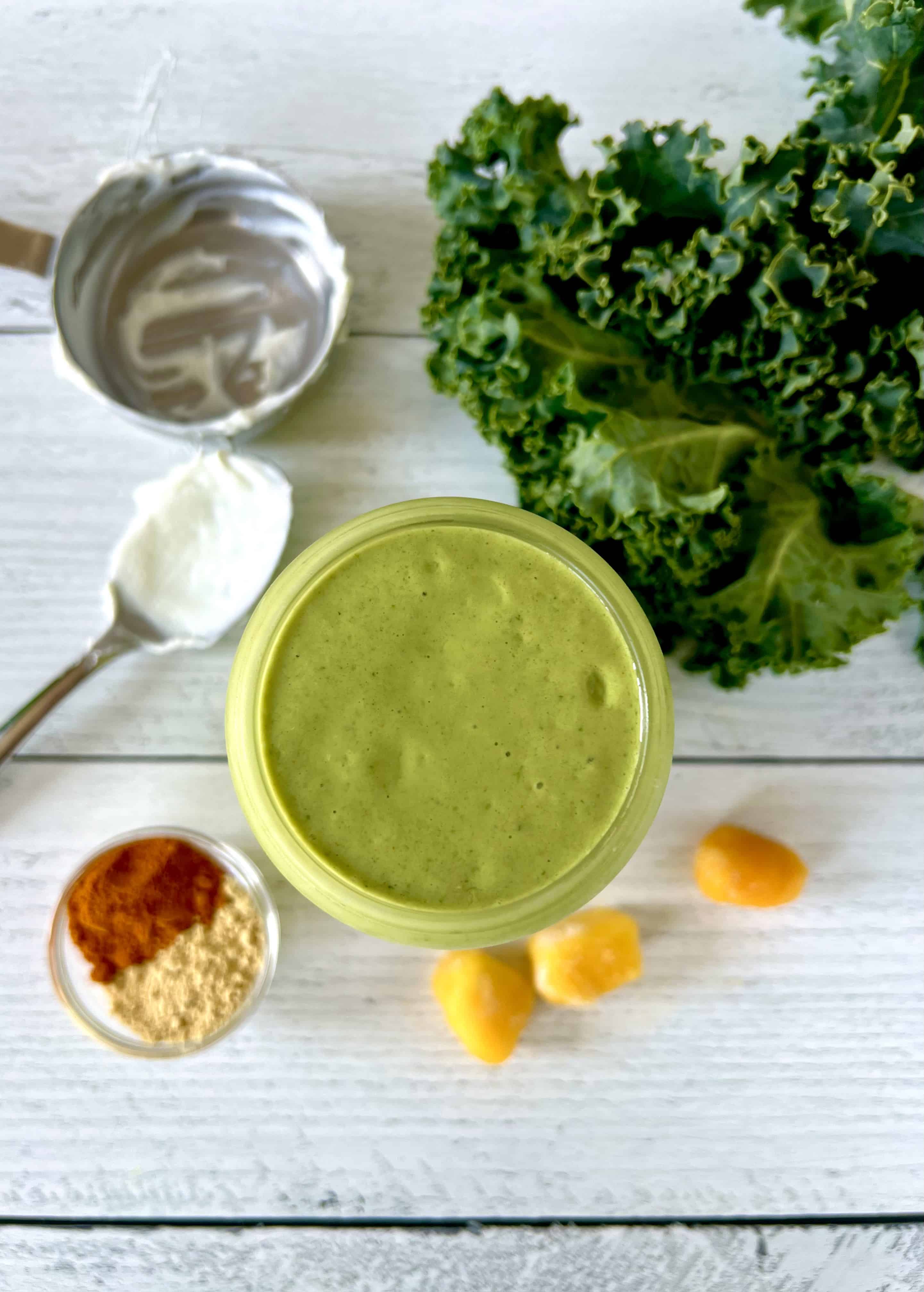 A vegan green smoothie in a glass on a white wooden table next to frozen mango chunks, kale leaves, a measuring cup, a big spoon with yogurt on it and a small bowl of spices.