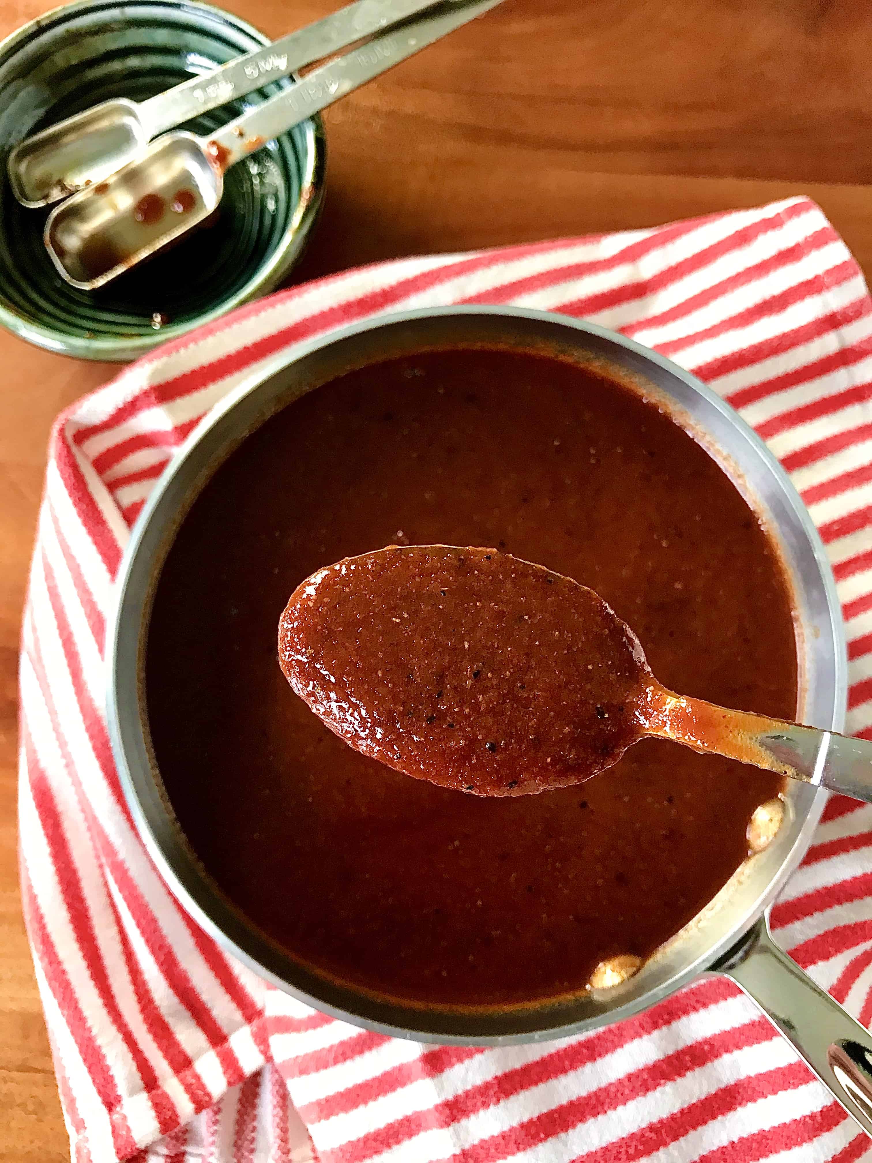 Gluten-free bbq sauce in a small pan on a red and white striped towel with a spoon over the pan holding some sauce.